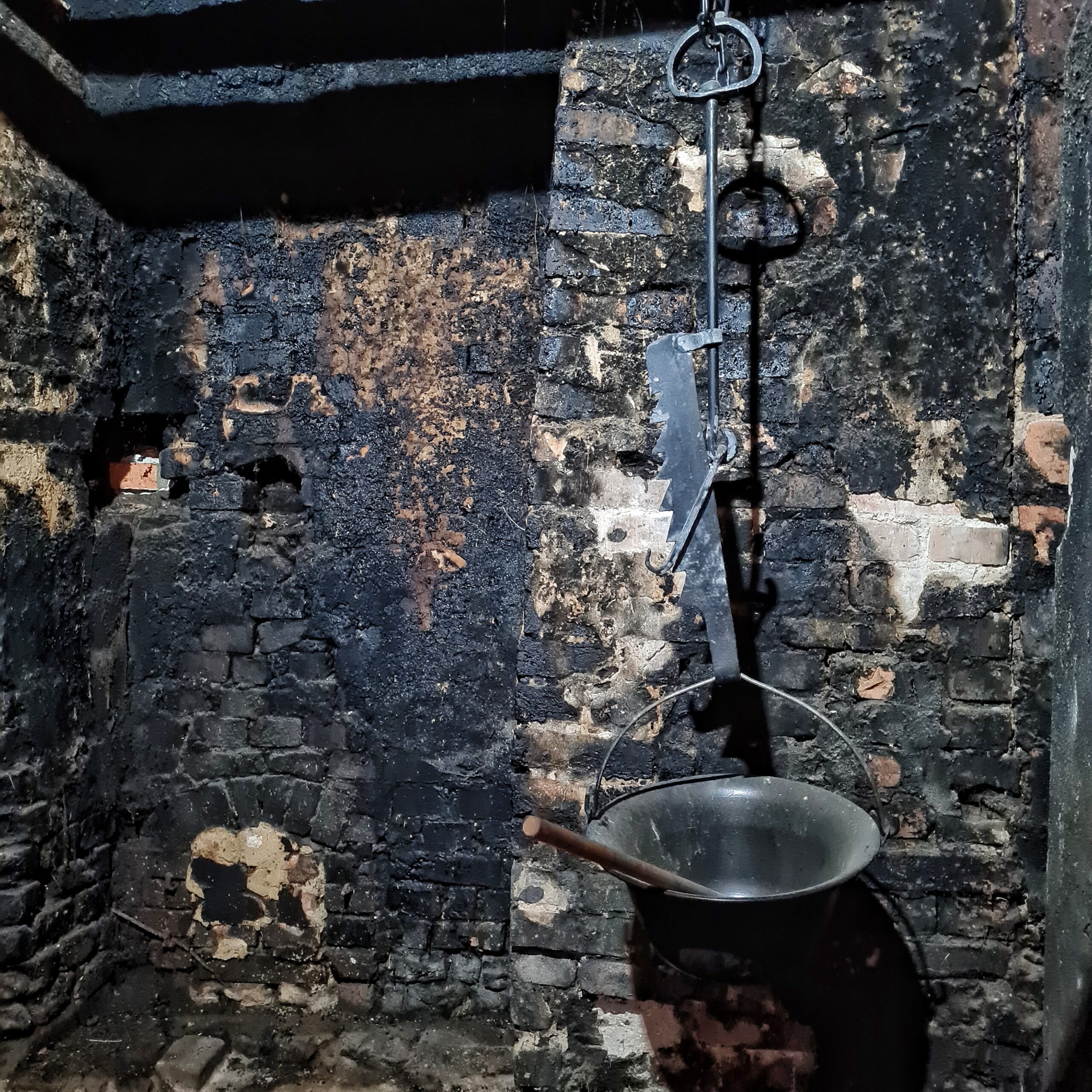 Eine rußgeschwärzte Mauer. Von oben hängt an einer Kette ein Topf herab. Darin ein Holzstiel.