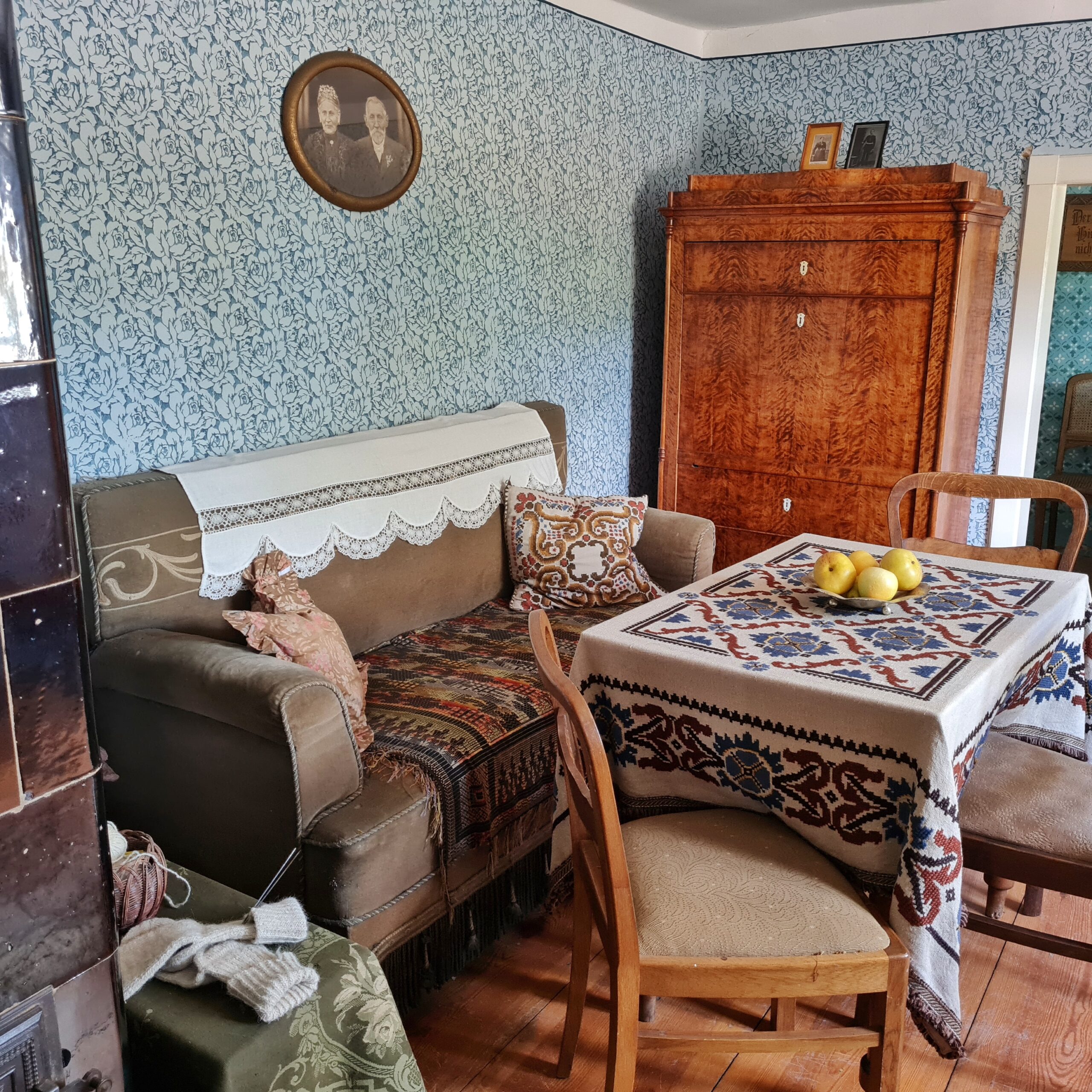 Eine alte Wohnstube mit blaugemusterter Tapete. An der linken Wand ein altes Sofa. Über der Rückenlehne hängt ein Spitzendeckchen. Auf der Sitzfläche eine bunte Decke und links und rechts gemusterte Kissen. Darüber ein rundes gerahmtes Schwarz-Weiß-Foto mit einer alten Frau und einem alten Mann. An der hinteren Wand ein Holzschrank. Darauf stehen zwei Fotos. Vorne rechts ein Tisch mit einer gemusterten Tischdecke und drei Holzstühle mit Plsterung. Auf dem Tisch steht eine Schale mit vier Äpfeln.