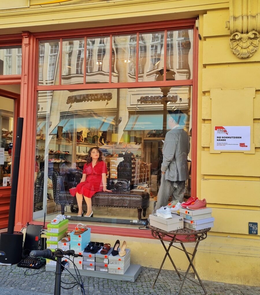 Ein Schuhladen in einem gelben Gebäude mit Verzierungen. Das Schaufenster ist von rötlich angestrichenem Holz umrandet. Oben sind Sprossen. Vor dem Schaufenster steht ein kleiner Tisch mit Schuhen darauf und danben  auf dem Boden Schuhkartons, auf denen Schuhe stehen. Daneben eine Lautsprecheranlage. Im Schaufenster stitzt eine Schuapoelerin im roten Kleid auf einem alten Ledersofa. Ein Schauspielerin einem hellgrauen Anzug steht seitlich von ihr. Die beiden reden miteinander. An der Wand neben dem Schaufenster hängt ein Schild, auf dem steht: Schauspielfenster. Die schmutzigen Hände. Von Jean-Paul Sartre. mit Guido Lambrecht und Mascha Schneider.Ab 14. Oktober 2022. Hans Otto Theater.