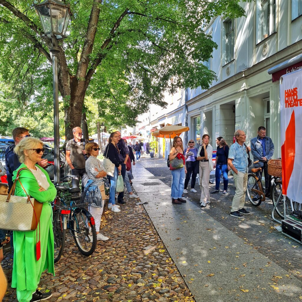 Ein breiter Bürgersteig, teilweise mit Kopfsteinpflaster. Er wird von Bäumen überschattet. Rechts eine Reihe von weißen Gebäuden. Auf dem Bürgersteig stehen mehrere Menschen und schauen interessiert auf etwas, was auf dem Foto nicht zu sehen ist.