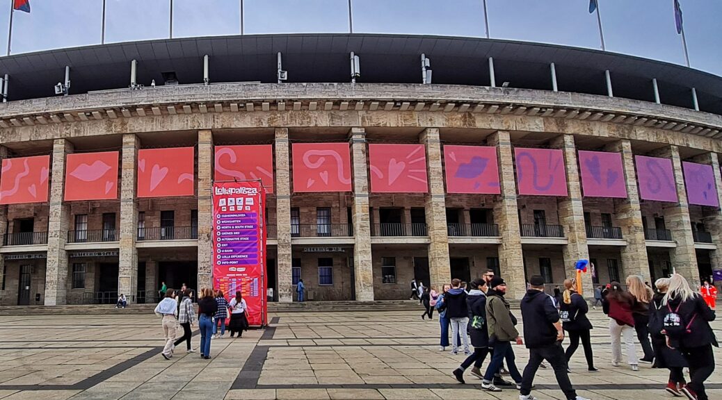 Das Berliner Olympiastadion von außen. Oben auf dem Gebäude bunte Fahnen, in der Mitte große Plakate mit Symbolen. Auf der freien gepflasterten Fläche laufen einige Menschen. Außerdem steht dort ein großer Aufsteller, audf dem oben Lollapalooza steht. Darunter sind die Logos von Sponsoren. Darunter werden mit Richtungspfeilen angezeigt: Fashionpalooza, Weingarten, North & South Stage, Grüner Kiez, Alternative Stage, Exit, U-Bahn, Lolla Experience, Info Point. Darunter das Line Up der Musiker*innen und Bands.
