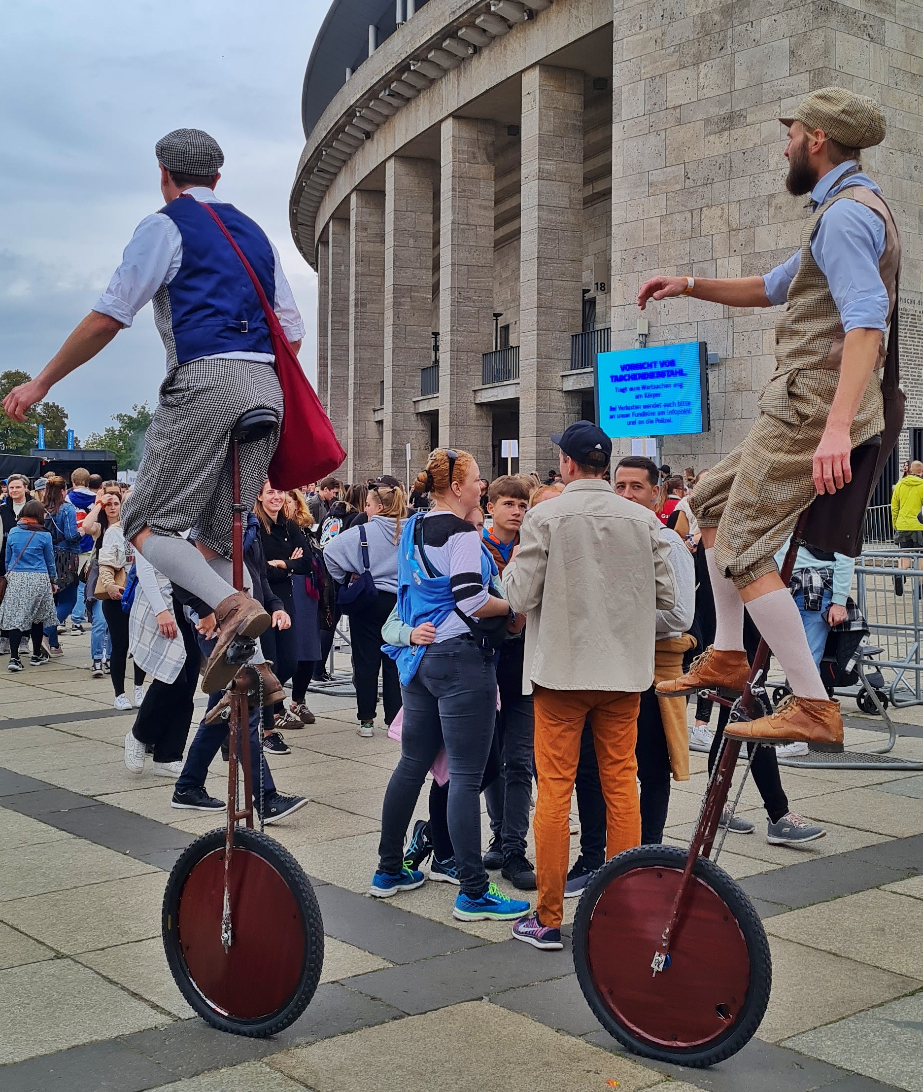 Vor dem Olympiastadion stehen Gruppen von Menschen und unterhalten sich. Zwei Einradfahrer in Knickerbocker, Westen und mit Mützen fahren durch.