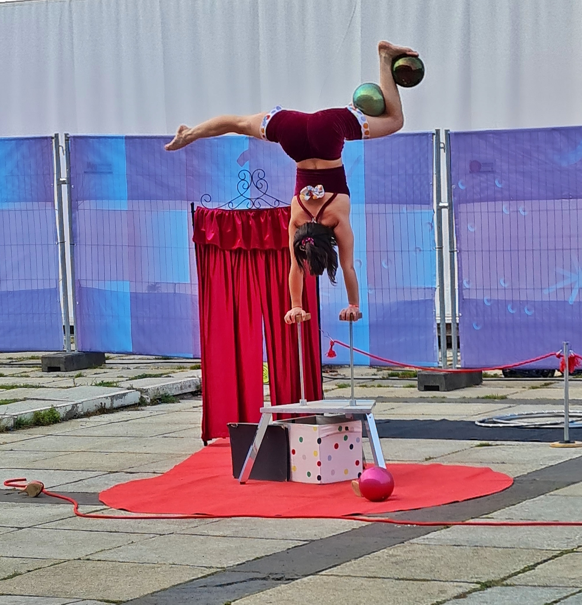 Ein rotes Tuch auf eine gepflasterten Fläche. Darauf ein Metallgestell mit zwei Stützen nach oben. Eine Frau in kurzen weinroten Hosen und einem weinroten Top hat ihre Hände darauf gestellt. Ihr rechtes Bein ist zur Seite ausgestreckt. Ihr linkes Bein ist angewinkelt. Zwischen Knie und Oberschenkel und zwischen Unterschenkel und Fuß ist jeweils ein glänzender Ball geklemmt.Dahinter ein Paravent mit rotem Stoff und dahinter ein Bauzaun.