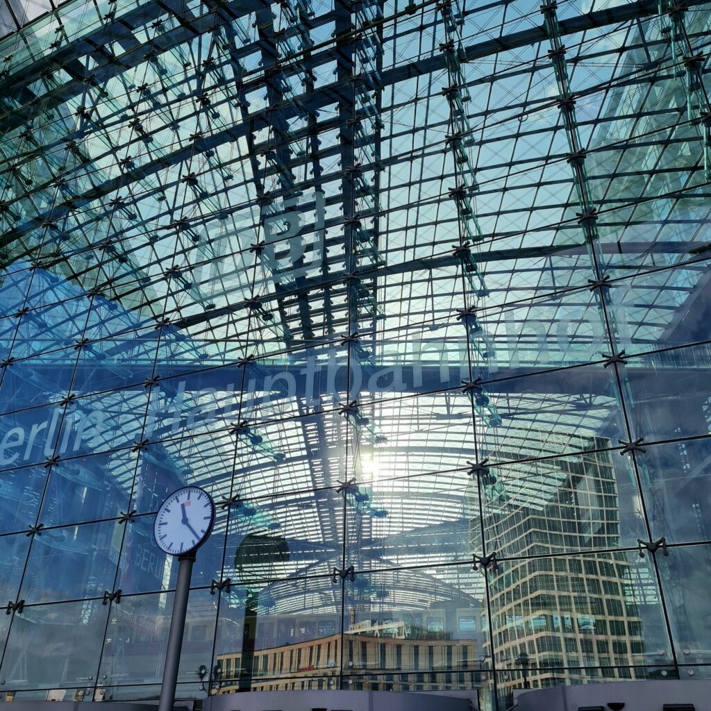 Der Berliner Hauptbahnhof von aussen fotografiert. Die Aufnahme wurde vom Europaplatz aus gemacht. Das Gebäude besteht aus Stahl und Glas, in dem sich gegenüberliegende Gebäude spiegeln. Vorne ist eine Bahnhofsuhr, die die Uhrzeit 22 Minuten nach 11 zeigt.