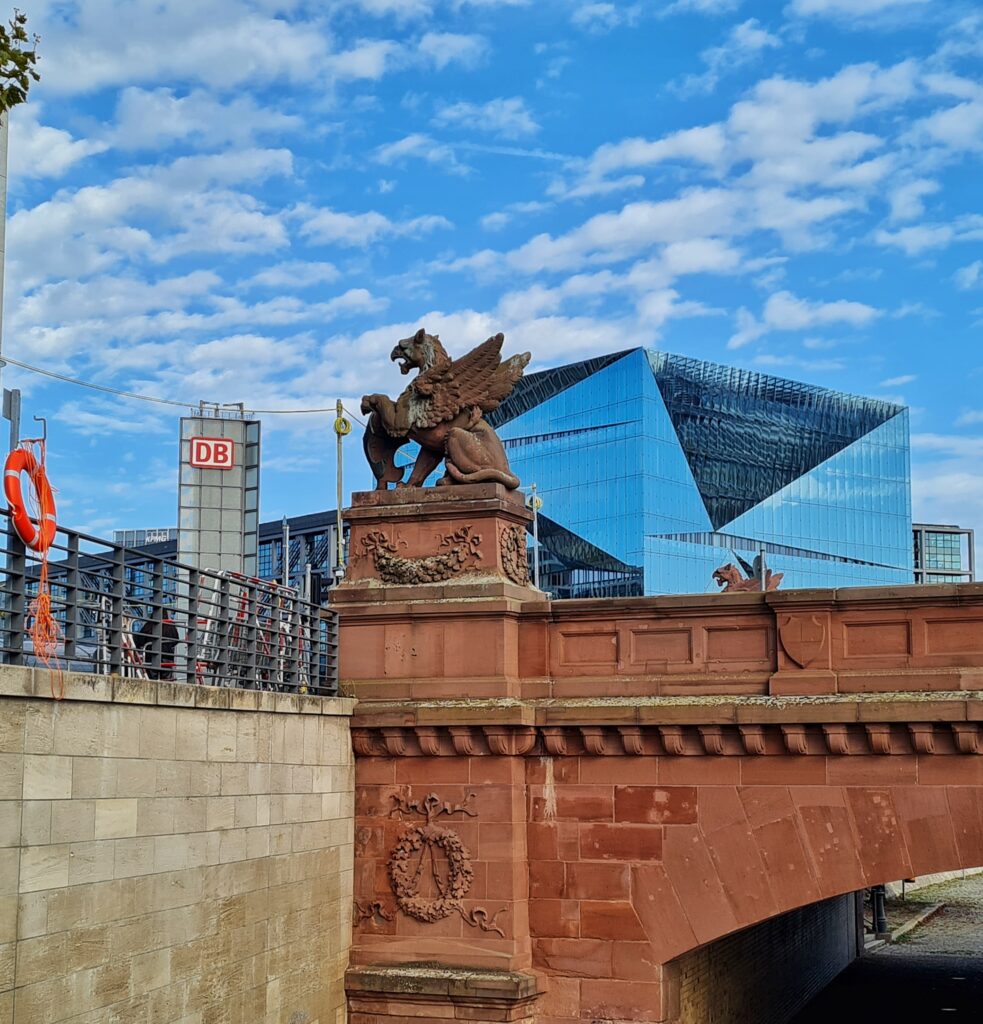 Blick von unten auf das Ende einer rötlichen Brücke mit einem geflügelten Löwen auf der Aussenseite. Dahinter der Cube Berlin. Daneben ein Turm mit der Aufschrift DB, und im Hintergrund der Hauptbahnhof.