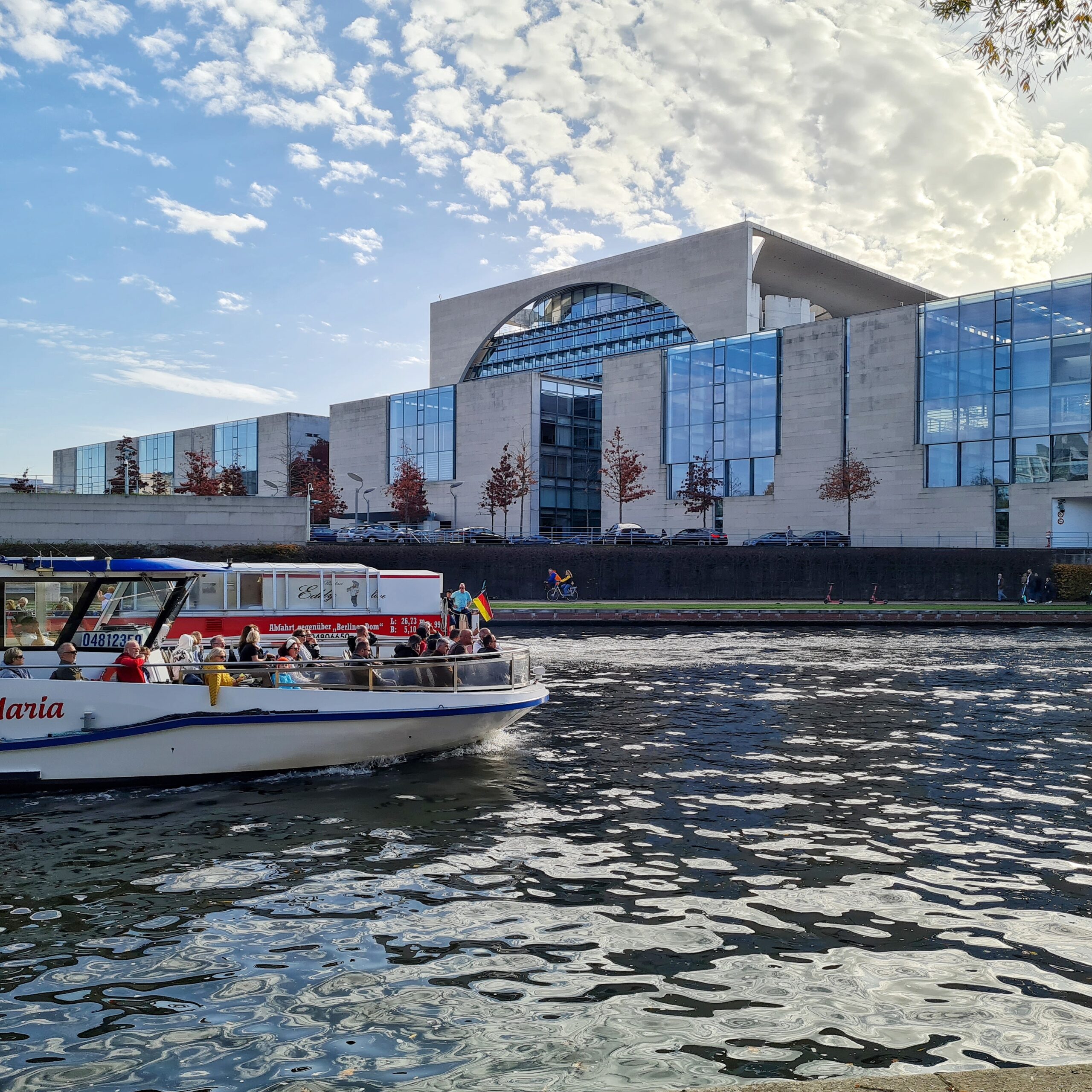 Blick über die Spree auf das Bubdeskanzleramt. Das hohe Hauptgebäude ist von der Seite zu sehen. Davor die Nebengebäude mit weiteren Büros. Davor stehen einige kleinere Bäume, und es parken mehrere schwarze Limousinen. Auf dem Weg davor laufen und radeln Menschen. Auf der Spree davor zwei Ausflugsschiffe. Von dem hinteren, das nach links fährt, sieht man den geschlossenen Heckbereich. Von dem anderen, das nach rechts fährt, den vorderen Teil, in dem Menschen im Freien sitzen.