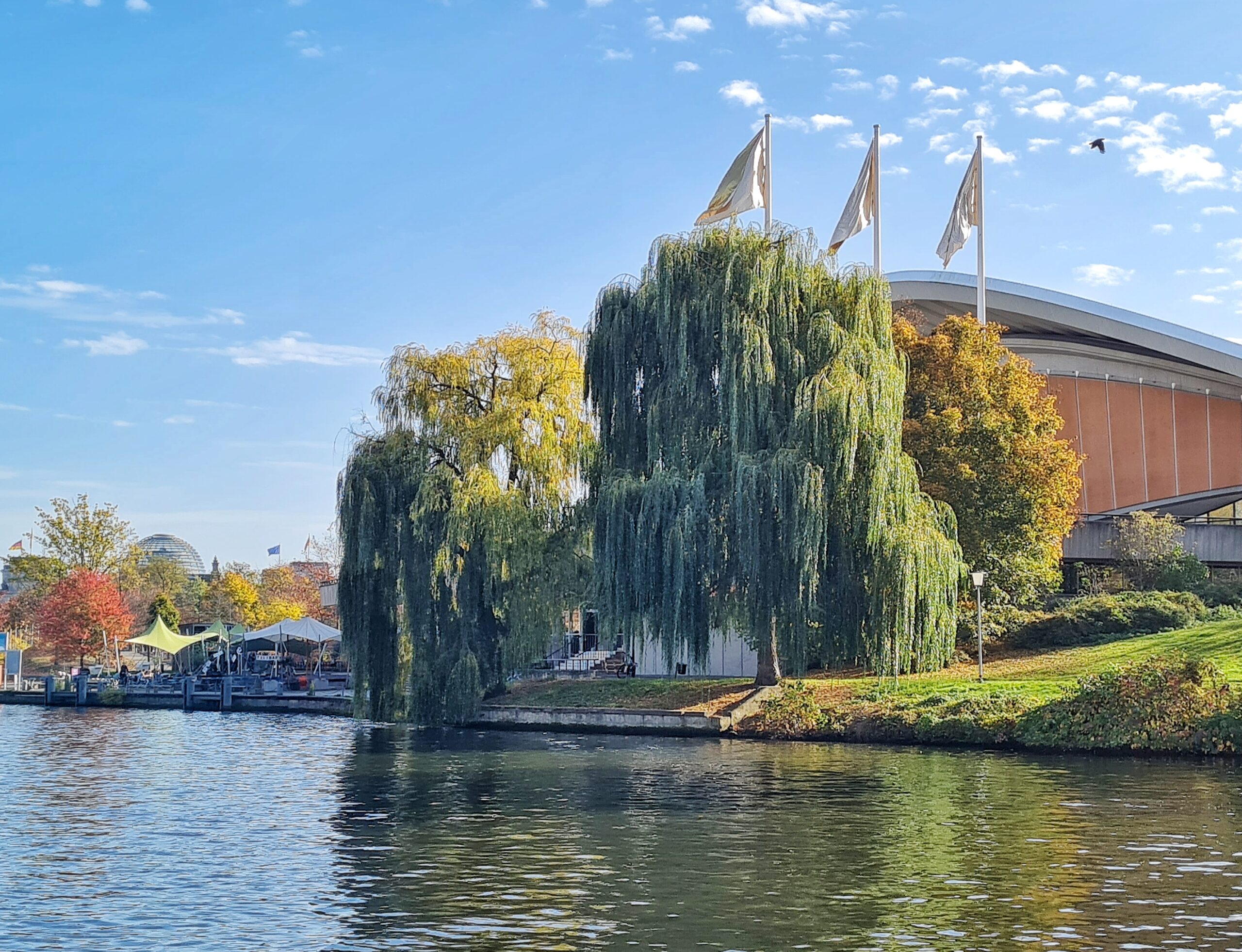 Blick über den Fluß auf das Haus der Kulturen der Welt, das größtenteils von Bäumen am Ufer verdeckt ist. Oben drei hohe Flaggen. Links liegen Sportboote.Dort sind auch Pavillons zu erkennen. Links im Hintergrund eine Glaskuppel.
