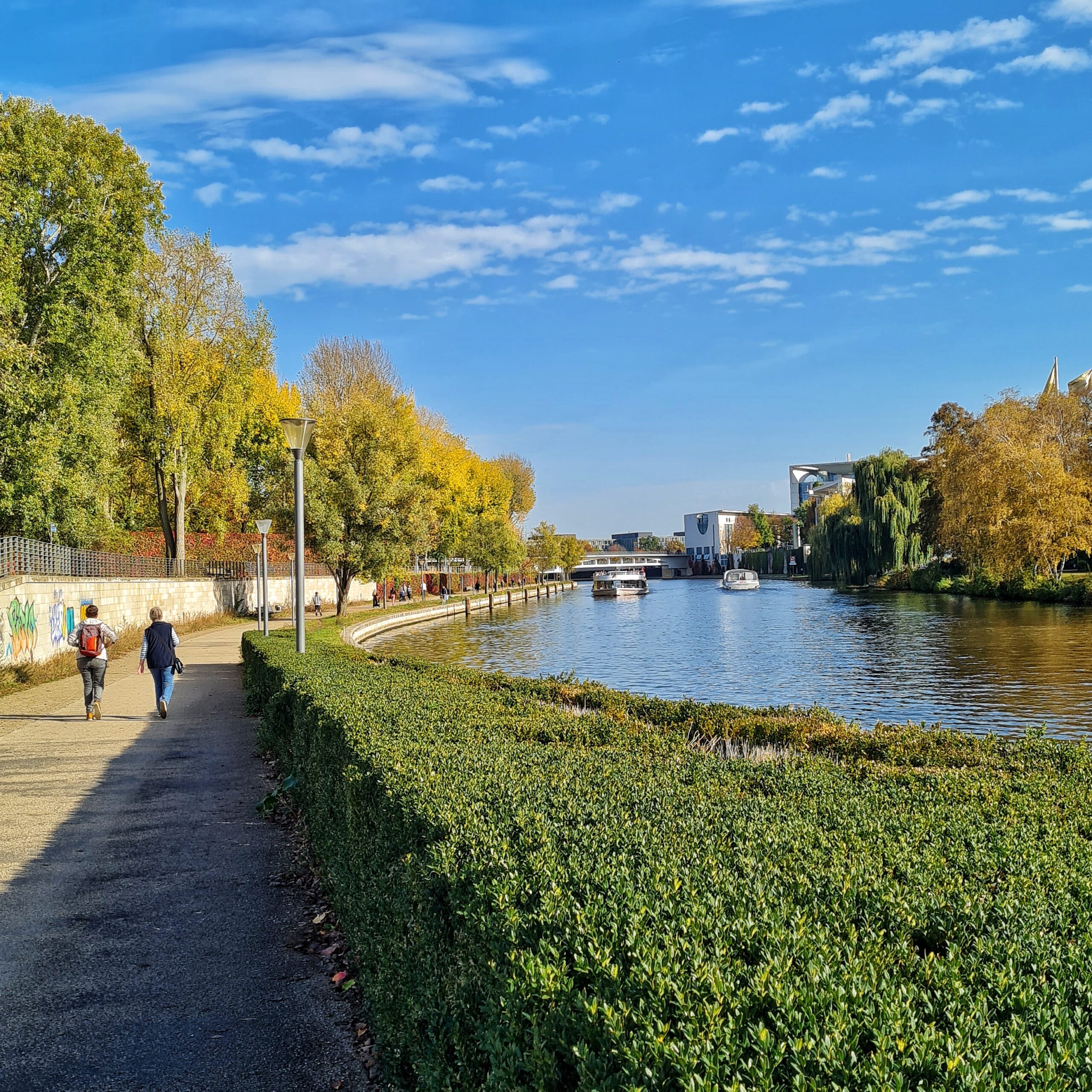 ein Gehweg entlang eines Flusses. Im vorderen Teil des Bildes ist zwischen Weg und Fluß eine gepflegte niedrige Hecke, im hinteren Teil eines Grasfläche mit Bäumen darauf, Auf dem Weg sieht man zwei Frauen von hinten. Viele Bäume auf beiden Ufern. Auf dem Fluß im Hintergrund zwei Ausflugsschiffe, die in unterschiedliche Richtungen fahren.