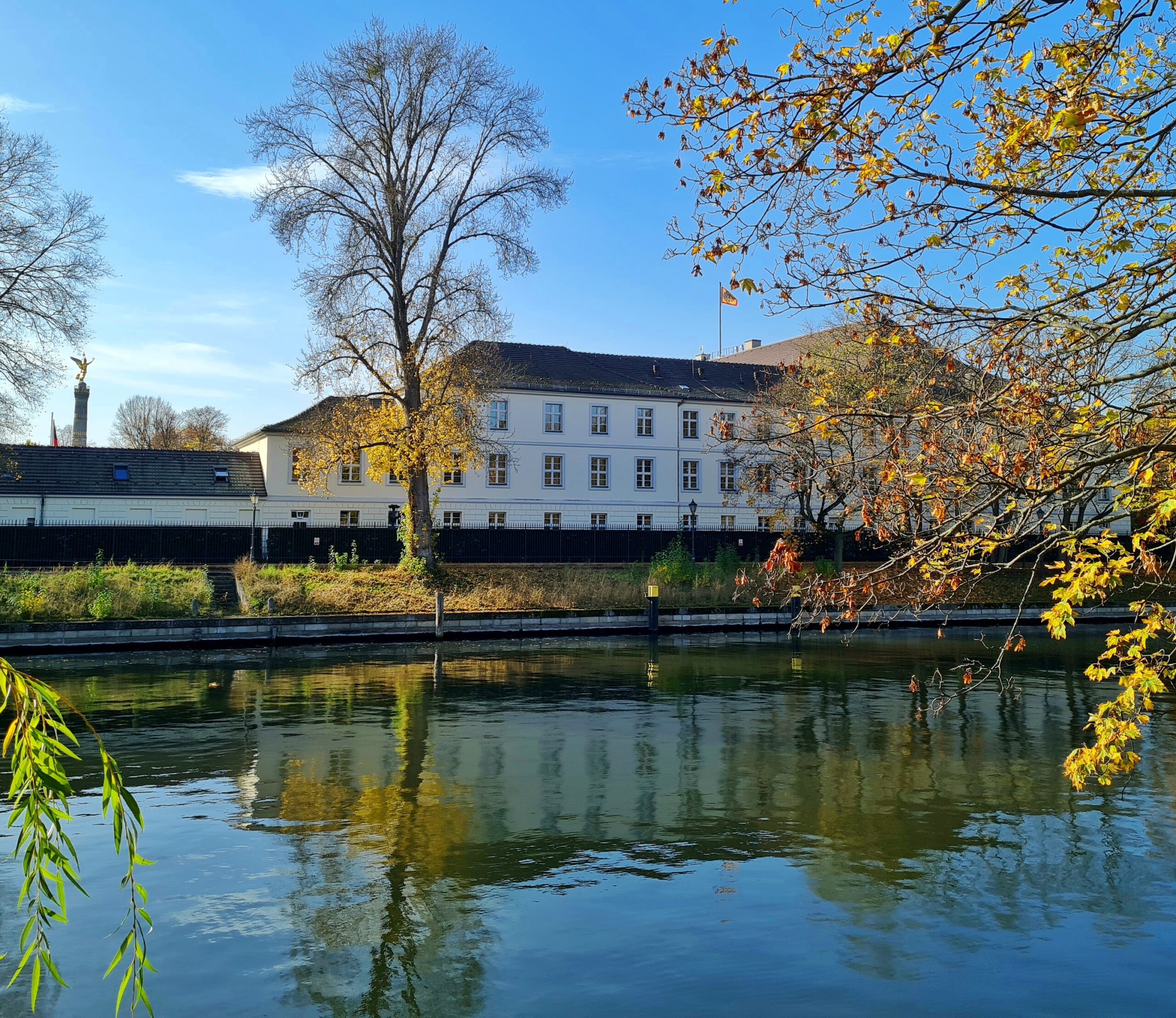 Blick über einen Fluß auf die Seitenansicht des Schloß Bellevie, teilweise verdeckt von gelblichen Bäumen, die an diesem Ufer stehen. Links im Hintergrund, sehr klein, die Siegessäule.