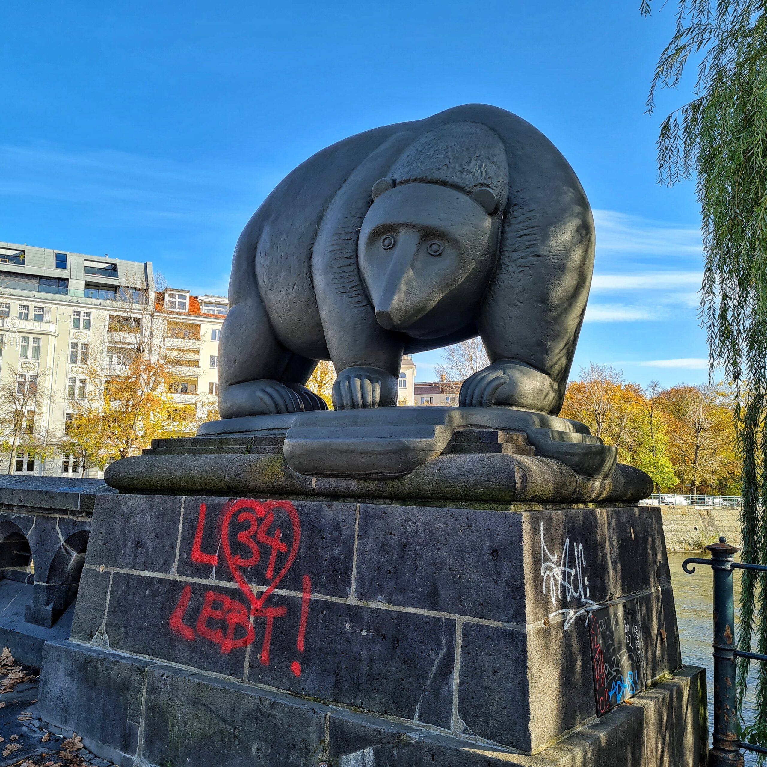 Eine rundliche Bärenfigur, mit allen ver Pfoten auf einem dunklen Steinsockel stehend. Auf den Sockel ist mit roter Fahrbe gesprüht: L 34 lebt. Um die 34 ist ein rotes Herz gesprüht. Im Hintergrund ein Fluß und auf dem anderen Ufer Grbäude und Bäume.