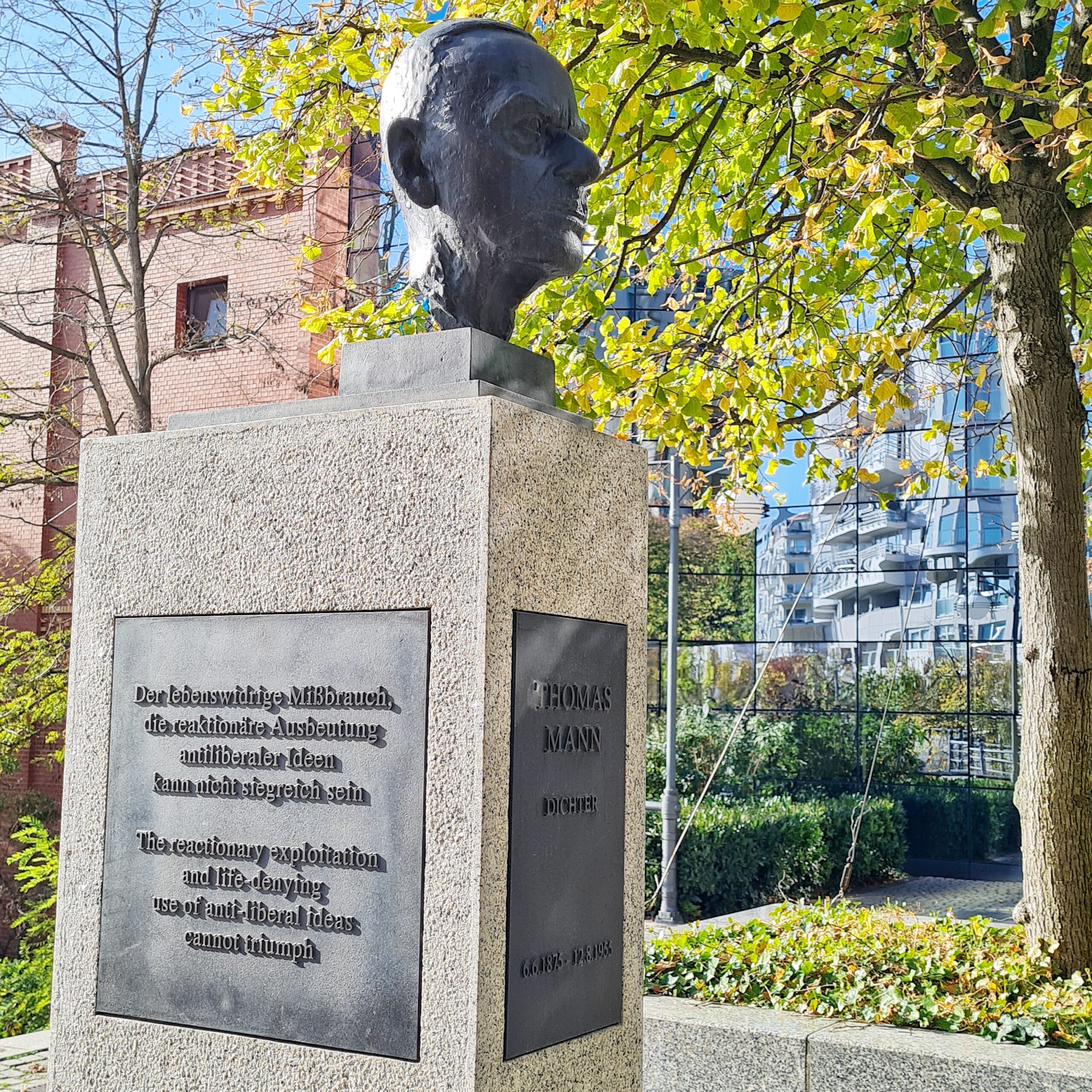 Ein hoher Strinsockel mit dem Metallkopf eines Mannes darauf. Vorne am Sockel steht auf einer Metallplatte: Thomas Mann, Dichter, 6.6.1875 - 12.8.1955. Auf einer seitlichen Metallplatte steht: Der lebenswidrige Mißbrauch, die reaktionäre Ausbeutung antiliberaler Ideen kann nicht siegreich sein. Im Hintergrund ein Backsteingebäude, an das sich ein Glasgeböäde anschließt.