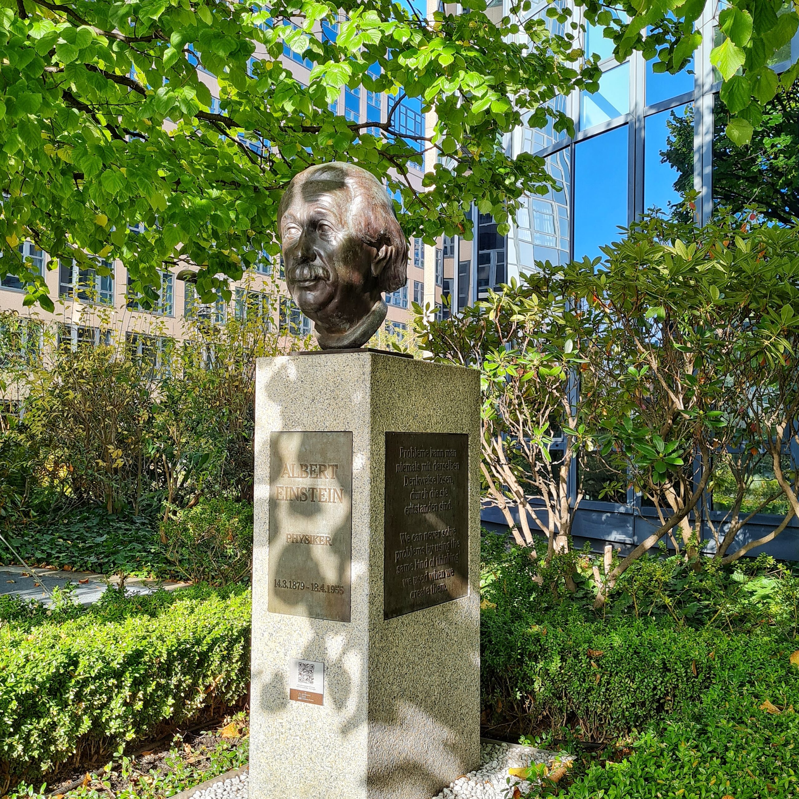 Bescbattet von Bäumen auf einem hohen Steinsockel der Metallkopf eines Mannes. Auf dem Sockel eine Metallplatte mit der Aufschrift: Albert Einstein, Physiker, 14.3.1879 - 18.4.1955. Auf der seitlichen Metallplatte steht: Probleme kann man niemals mit derselben Denkweise lösen, durch die sie entstanden sind.