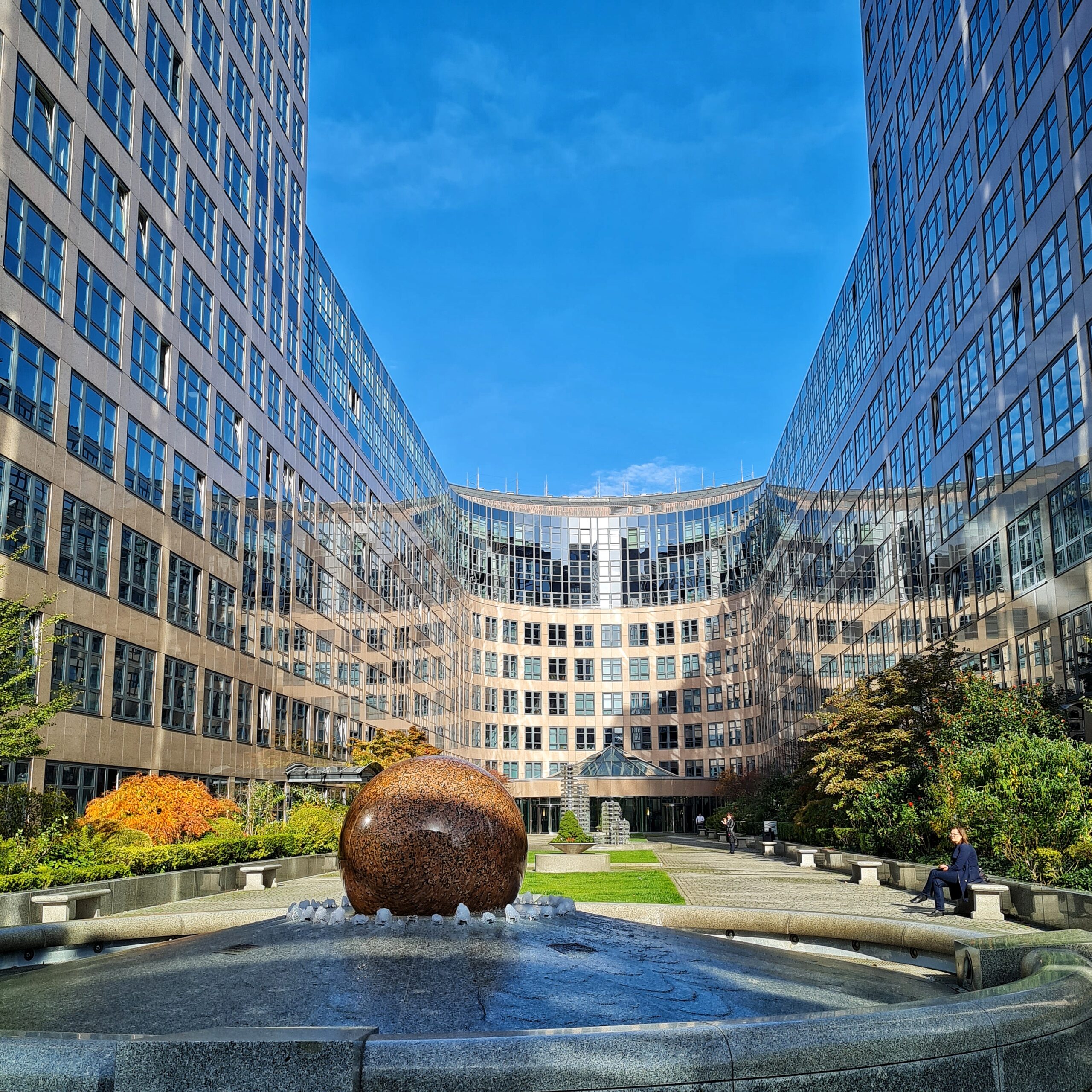 Um einen Innenhof eine geschlossene Fassade eines mehrstöckigen Grbäudes mit viel Glas. Es ist U-förmig. Vorne, das heißt am offenen Teil des U, ein großer Brunnen mit einer großen bräunlichen Steinkugel darin.