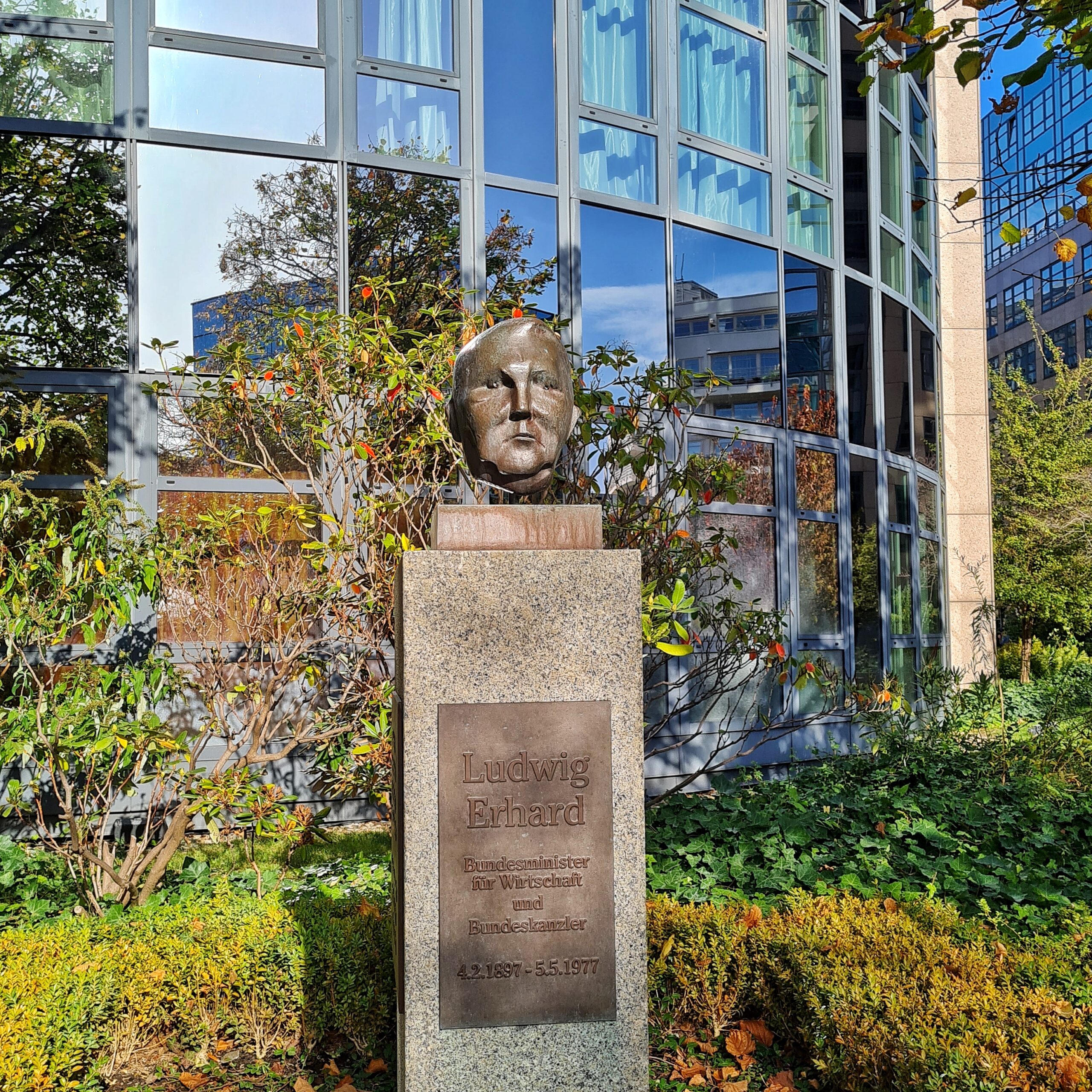 Vor einem Glasgebäude ein hoher Steinsockel, auf dem der Metallkopf eines Mannes steht. auf der Metalltafel am Sockel steht: Ludwig Erhard, Bundesminister für Wirtschaft und Bundeskanzler, 4.2.1897 - 5.5.1977.