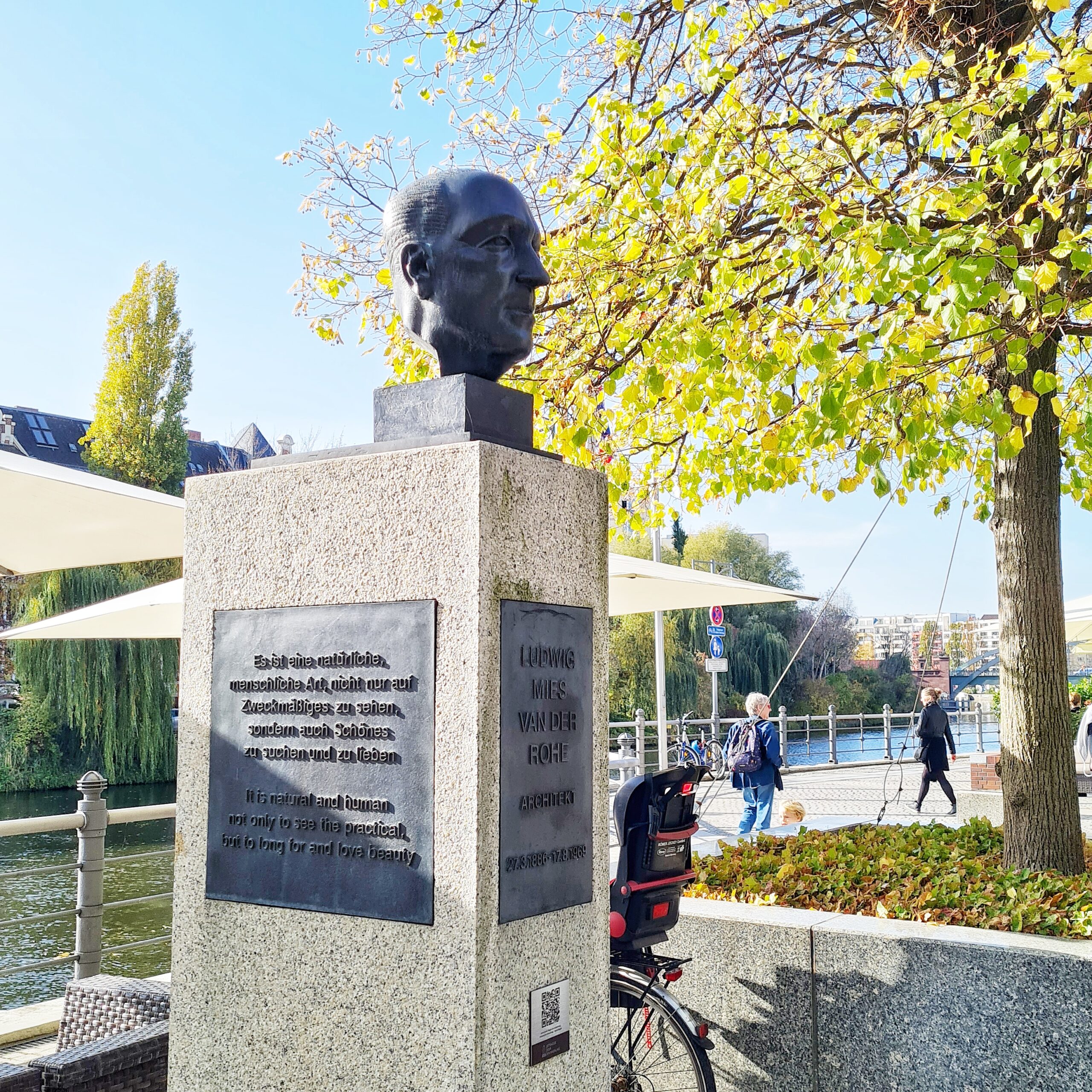 Ein Männerkopf aus Metall auf einem hohen Steinsockel. Die Aufnahme ist halb seitlich gemacht worden, so dass man die Vorderseite und eine Seite des Steinsockels sehen kann. Auf der Vorderseite ein Metallschild mit der Aufschrift: Ludwig Mies van der Rohe, Architekt, 27.31886 - 17.9.1969. Auf der Seite ebenfalls ein Metallschild mit der Aufschrift: Es ist eine natürliche, menschliche Art, icht nur auf Zweckmäßiges zu sehen, sondern auch Schönes zu suchen und zu lieben. Im Hintergerund ein Fluß und eine Promenade mit Pavillons, auf der einige Menschen gehen.