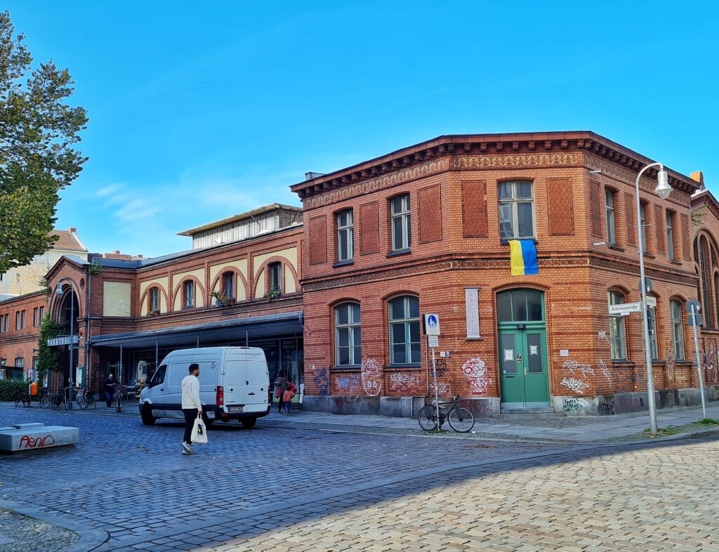 Ein zweistöckiges längliches Gebäude, weitgehend aus rötlichem Backstein. Der Eckteil hat eine grünliche Tür, über der eine Ukraine-Flagge hängt und mehrere Fenster. Links ist das Eingangsportal zu sehen. Dazwischen ist offenbar im Erdgeschoss ein Laden, der jedoch fast vollständig von einem weißen Lieferwagen verdeckt wird. Vor dem Gebäude laufende Menschen und parkende Fahrräder.