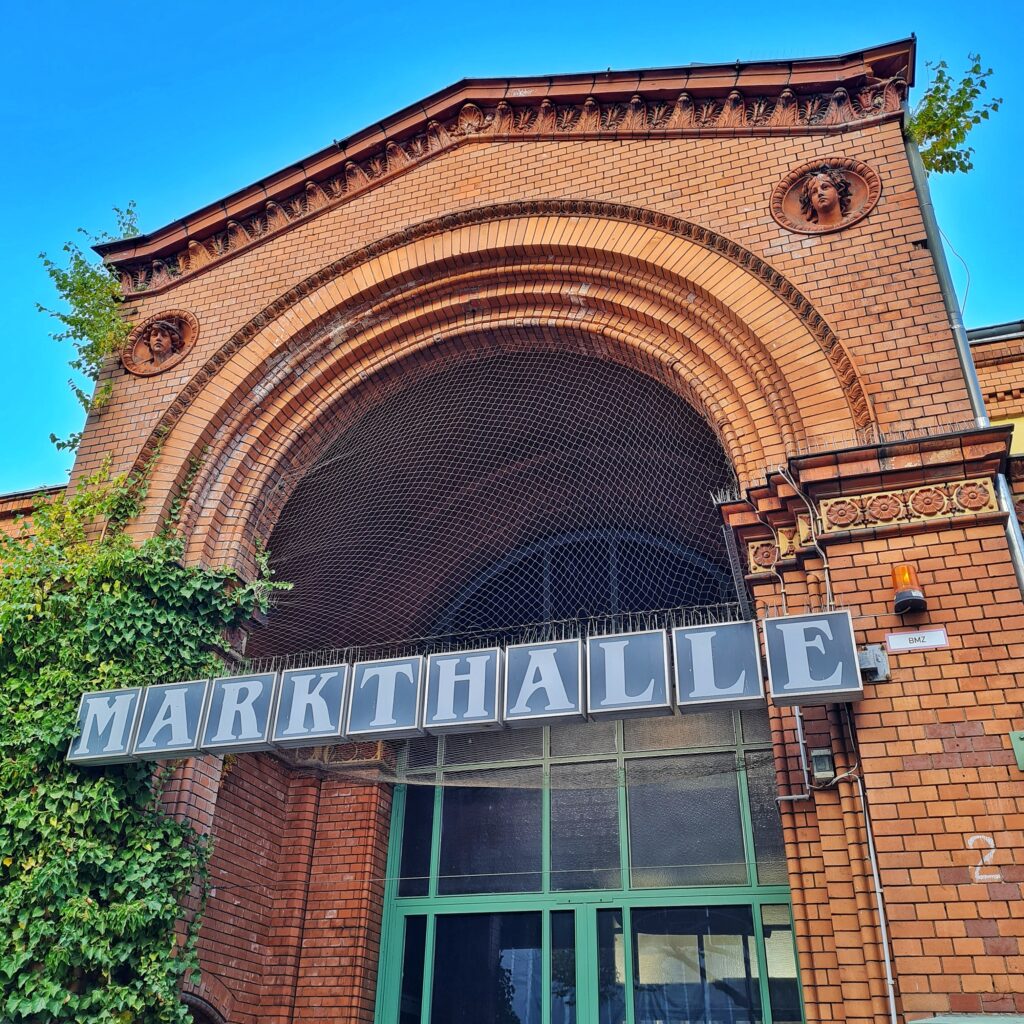 Das hohe Eingangsportal der Markthalle aus bräunlich-rötlichem Backstein. Oben Verzierungen, darunter zwei Menschenköpfe. Der obere Teil des Portals ist mit einem feinen Gitter versehen. Darunter hängen große Buchstaben, die das Wort "Markthalle" bilden.