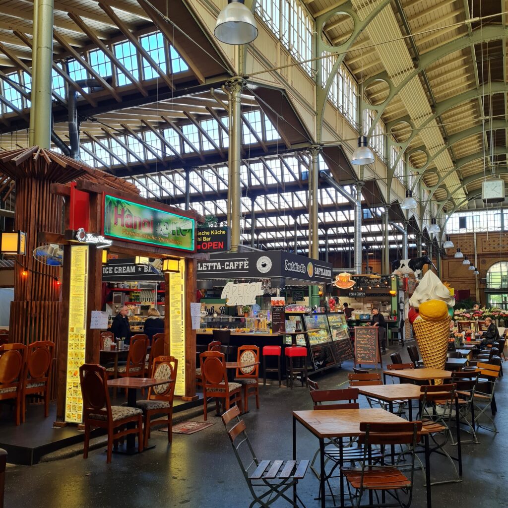 Blick in die Markthalle. Die Dachkonstruktion wirkt mit vielen Bögen leicht. In das Dach siind viele Fensterreihen eingelassen, so dass es in der Halle hell ist. Auf der rechtren Seite mehrere Buden und Restaurants: ein vietnamesisches Restsurant, ein Stand mit Kaffee und Eis sind im Vordergrund zu erkennen. Links mehrere kleine Tische mit Stühlen und die Figur einer großen Eiswaffel mit Eiskugeln darin.