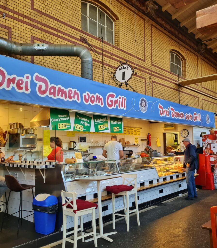 An der Wand der Markthalle (gelber Backstein mit dekorativen roten Linien dazwischen) ein Imbiss, in dem zwei Frauen und ein Mann arbeiten. An der Theke wartet ein Kunde. Über dem Imbiss ein hellblaues Banner mit der Aufschrift "Drei Damen vom Grill". Darunter hängen Schilden mit den Aufschriften: Riesen-Currywurst 2,70 Euro, Pommes Frites 2,20 Euro, Eintopf 4,00 Euro, Pott Kaffee 2,00 Euro.