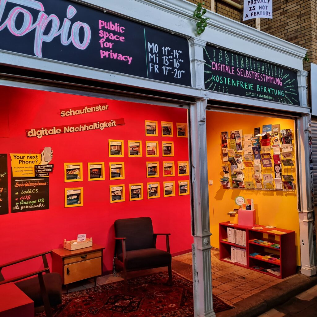 Ein Laden, nach vorne offen. Darüber steht: Topio. public space for privacy, Mo 11 - 14, Mi 13 - 16, FR 17 - 20, Digitale Selbstbestimmung, Kostenfreie Beratung. Auf dem Dach ein Schuld an einem Holzstock mit der Aufschrift: Privacy is not a Feature. Im Laden zwei Sessel mit einem niedrigen Schränkchen dazwischen, ein niedriges Regal und an den Wänden Fotos, Schilder und ein Regal mit Broschüren. An der breiteren Wand steht darüber: Schaufenster digitale Nachhaltigkeit.