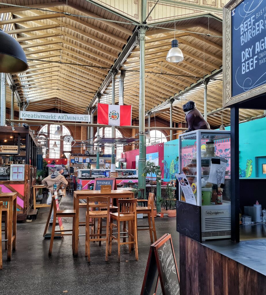 Rechts ein Teil des Tresens eines Burgerrestaurants. Auf ihm steht ein Glasbehaälter mit Getränken darin; darauf die Figur eines Mopses. Dahinter Stühel und Tische. Dahinter weitere Stände. Oben hängt ein Banner: Kreativmarkt ATminius 2. Daneben hängt eine Flagge. Ganz im Hintergrund hohe Fenster.