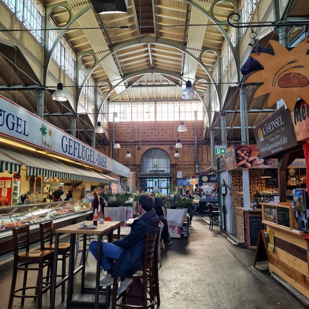 Ein Gang in der Markthalle, bei dem man gut die Bögen der Hallenkostruktion sehn kann. Das Licht kommt durcch eine durchgehende Fensterreihe unter der Decke. Links und rechts Marktstände, wo man Geflügel, Käse, Brot und ähnliches kaufen kann. In der Mitte ein paar Tische und Stühle. an einem Tisch sitzt ein Mann, der einen geleerten Teller und eine halbvolle kleine Falsche Cola vor sich hat. Dahinter ein Stand mit Blumen.