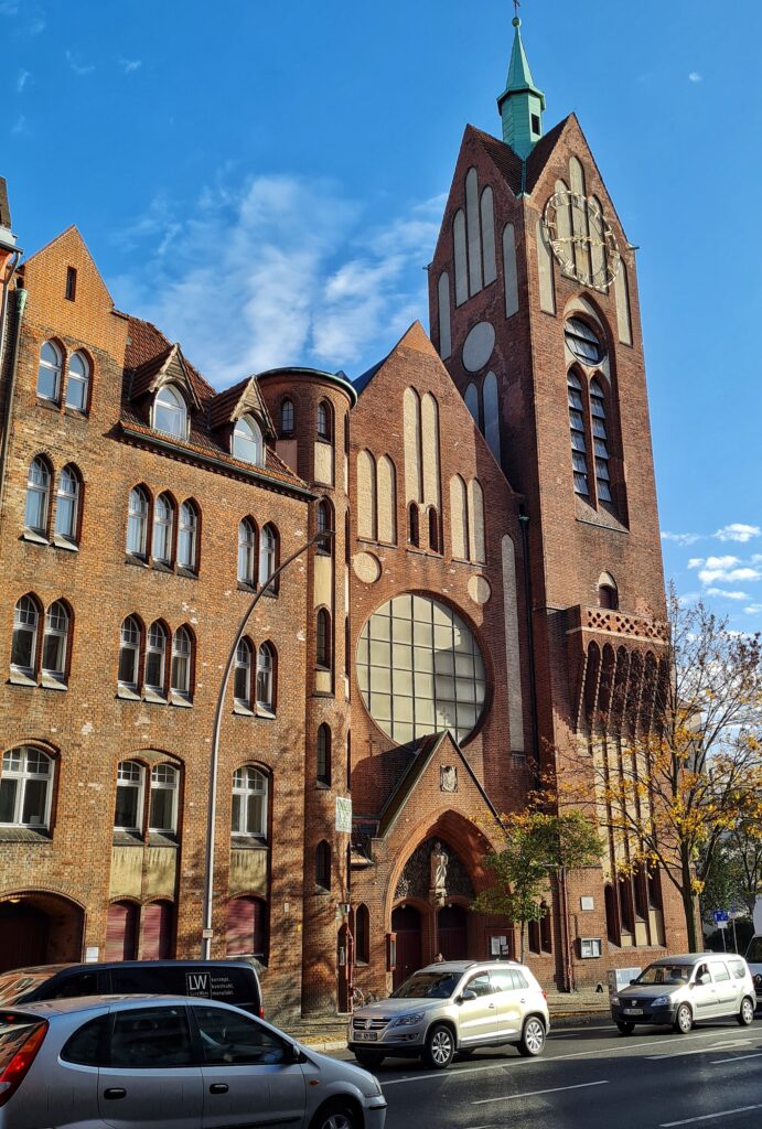 Eine mit Autos befahrene Straße. Auf der anderen Straßenseite neben einem mehrstöckigen Backsteingebäude eine Backsteinkirche. Sie hat einen schmalen Eingangsbereich und daneben einen hoch aufragenden viereckigen Kirchturm.