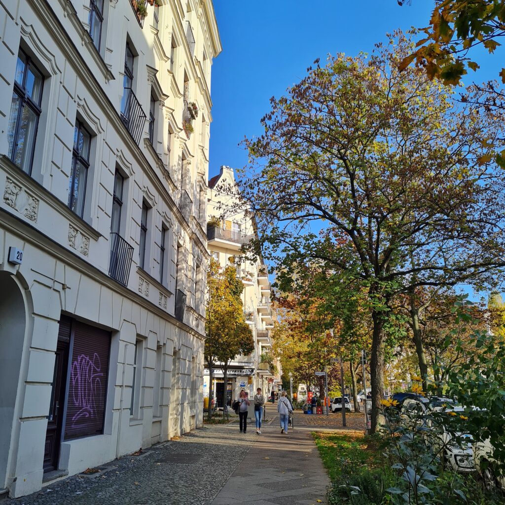 Ein breiter Gehweg, auf dem mehrere Menschen gehen. Links mehrstöckige weiße Wohnhäuser mit Verzierungen. Auf der anderen Seite des Weges eine lange Reihe von Bäumen auf einem Grünstreifen. Daneben parkende Autos.