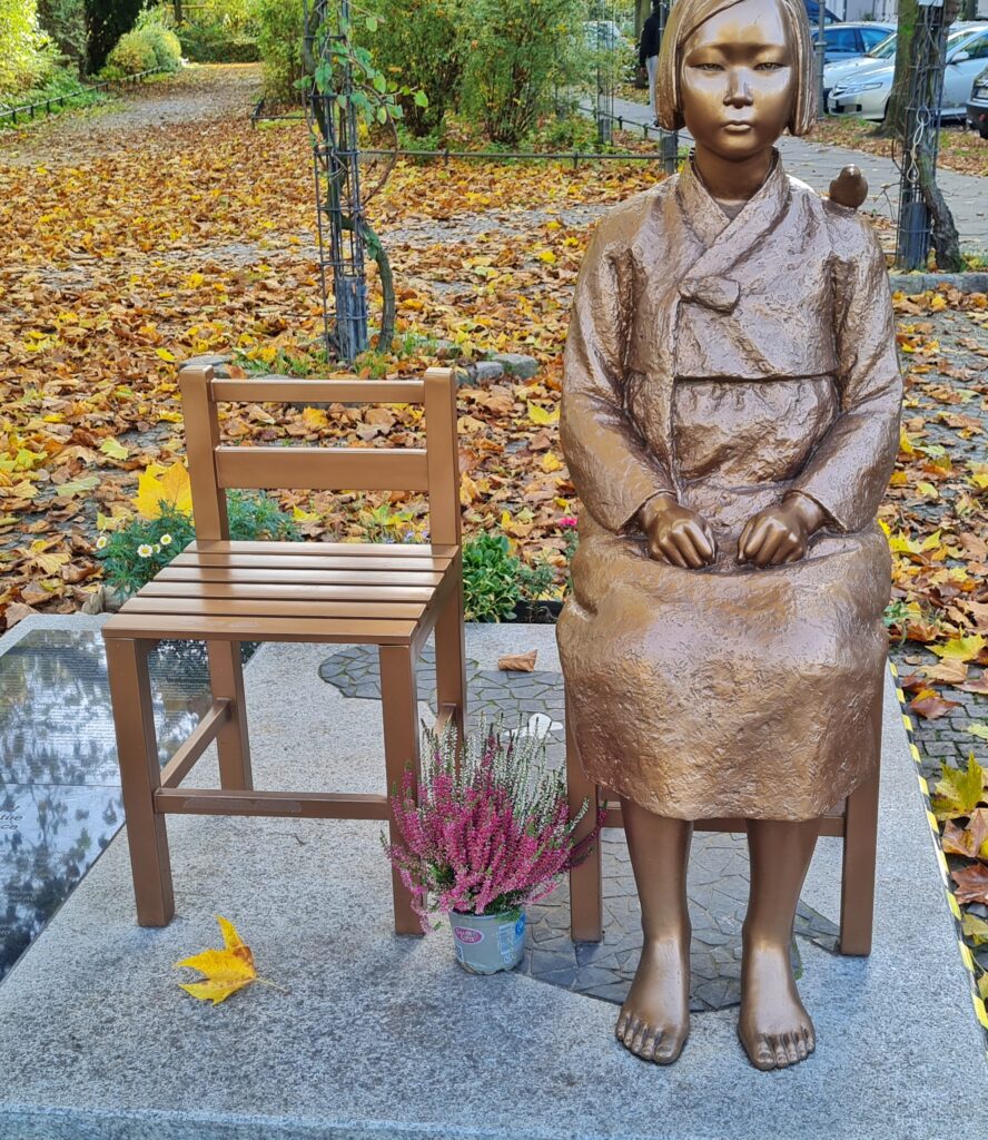 Eine Metallskulptur auf einer Bodenplatte. Zwei Stühle nebeneinander, einer leer. Auf dem anderen sitzt ein junges Mädchen in koreanischer Tracht. Sie ist barfuss. Ihre Hände liegen zusammengeballt in ihrem Schoß. Auf ihrer Schulter hockt ein kleiner Vogel. Zwischen den Stühlen steht ein kleiner Topf mit einer blühenden Pflanze. Im Hintergrund eine kleine Parkanlage mit Herbstlaub auf dem Boden und eine Straße mit parkenden Autos.