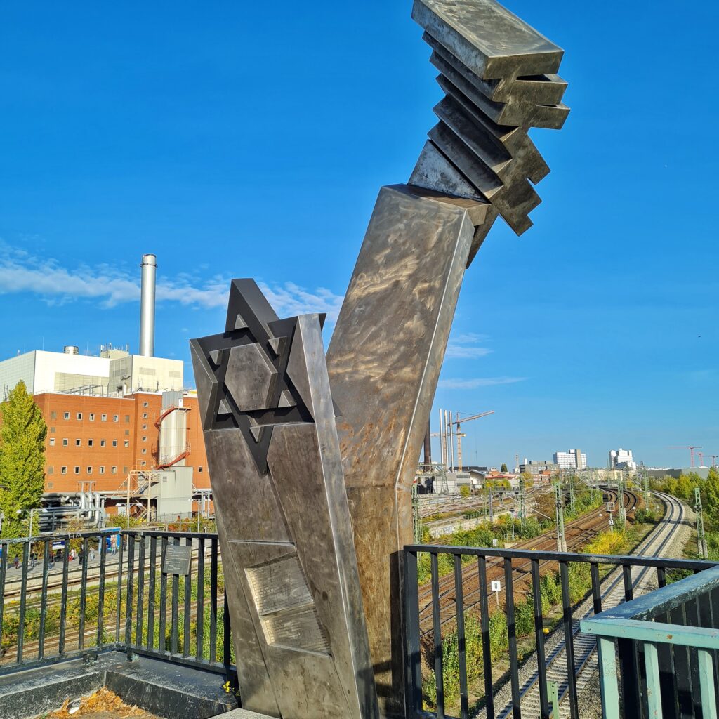 Eine Metallskulptur, bestehend aus zwei breiten Metallbändern. Der vordere, kürzere zeigt oben einen großen Judenstern. Unten ist Text eingraviert. Der hintere, höhere läuft oben in schräg aufgestapelten Metallplatten ausläuft. Im Hintergrund unten Bahngleise. Links unten ein Industriegebäude mit einem hohen weißen Schornstein.
