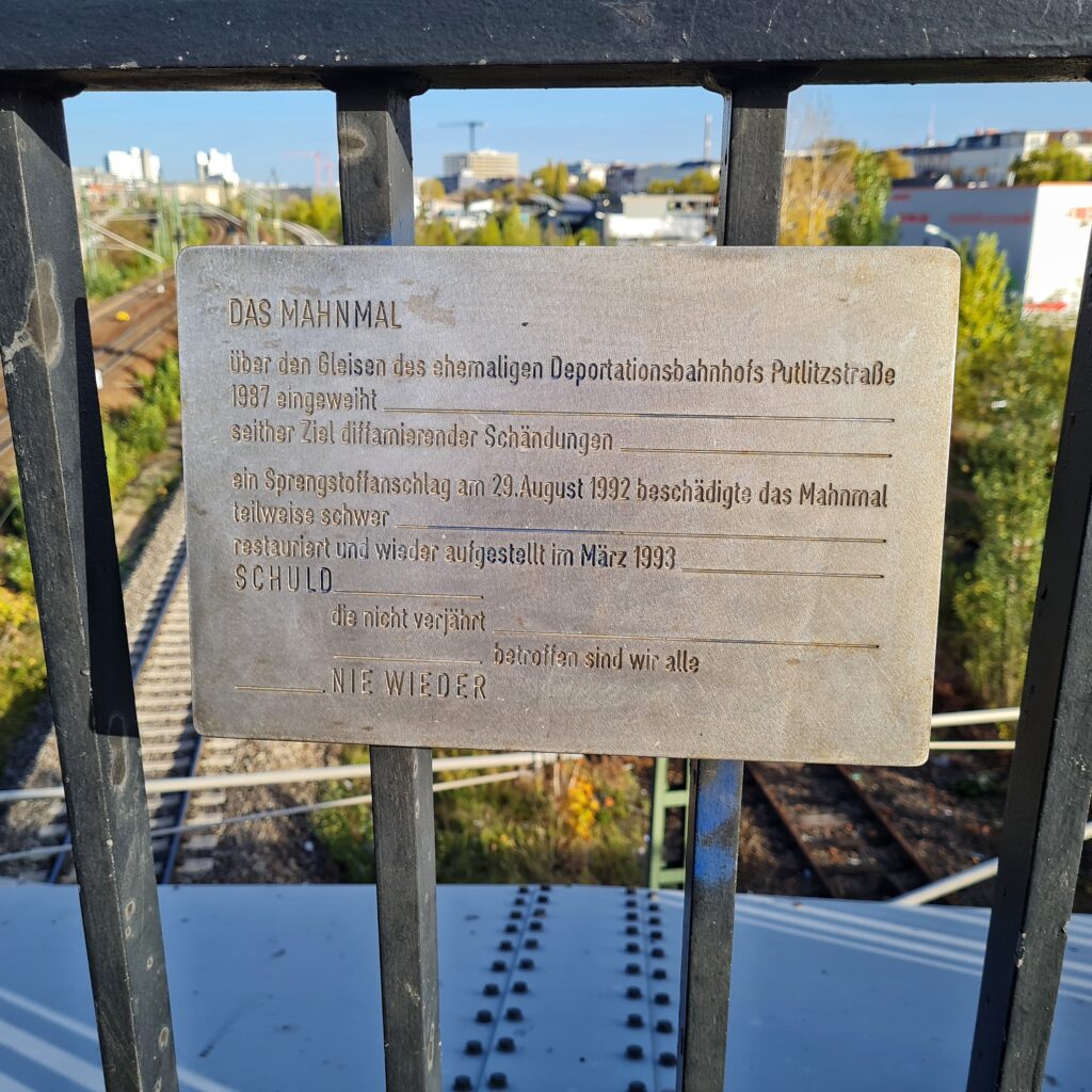 Ein Metallschild an einem Brückengeländer mit der Aufschrift: Das Mahnmal über den Gleisen des ehemaligen Deportationsbahnhofs Putlitzstraße, 1987 eingeweiht, seither Ziel diffamierender Schändungen, ein Sprengstoffanschlag am 29. August 1992 beschädigte das Mahnmal teilweise schwer, restauriert und wieder aufgestellt im März 1993, Schuld, die nicht vergeht, betroffen sind wir alle, Nie wieder. Im Hintergrund, etwas versschwommen, Gleise, hohe Gebäude und Bäume.