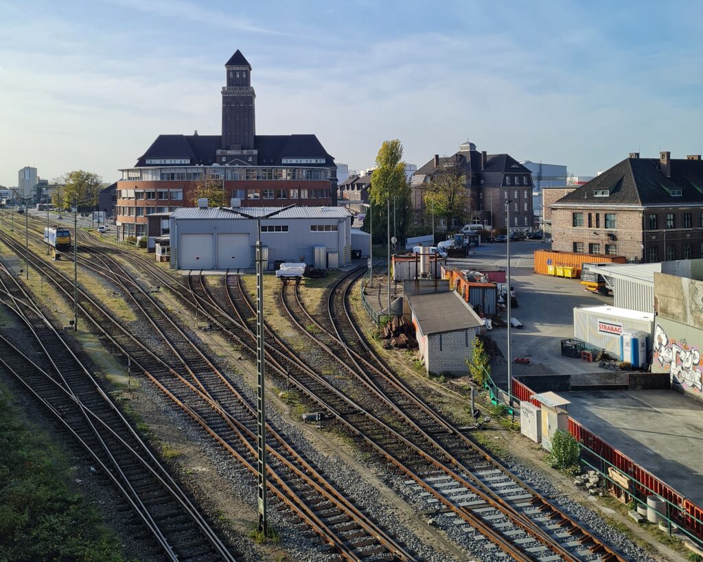 Blick von oben auf Gleise. Im Hintergrund verschiedene große Grbäude, eines davon mit einem hohen viereckigen Turm. Rechts Container und Schuppen.