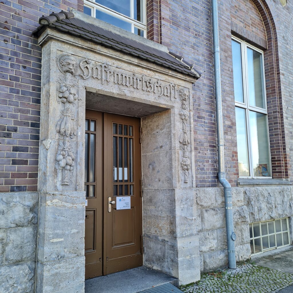 Die Eingangstür eines Backsteingebäudes aus bräunlichem Holz. Sie wird eingerahmt und überdacht von einem Steinportal, auf dem oben "Hafenwirtschaft" steht. Rechts und links sind Reliefe aus Stein, die oben einen Männerkopf und darunter Obst und Gemüse zeigen. Neben und oberhalb der Tür sind Fenster.