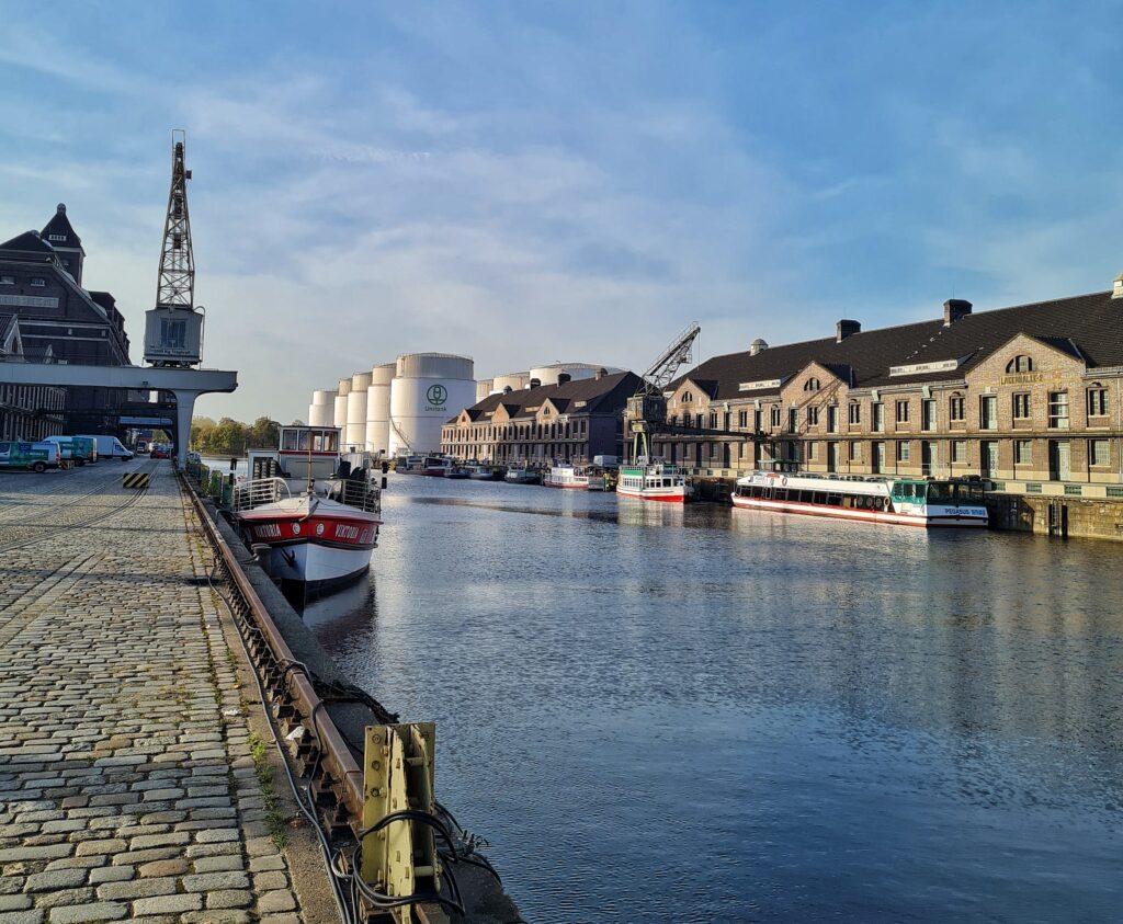 Aufnahme eines Hafenbeckens zur offenen Seite hin. Links ist ein Teil eines großen Backsteingebäudes zu sehen. Auf dem Kai ein grauer Kran. Im Wasser ein Ausflugsschiff. Auf der rechten Seite zwei große Lagerhallen und große, weiße Tanks. Am Kai liegen Ausflugsschiffe.