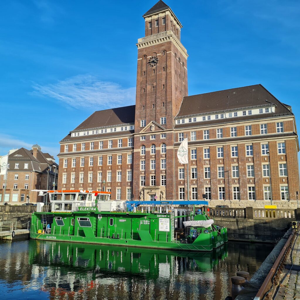 Am Ende des Hafenbeckens ein großes, altes Backsteingebäude mit einem hohen, quadratischen Turm, an dem ein Ziffernblatt angebracht ist. Das Gebäude hat viele weiße Sprossenfenster. Im Wasser davor ein grünes Metallboot mit dem Namen "Elektra".