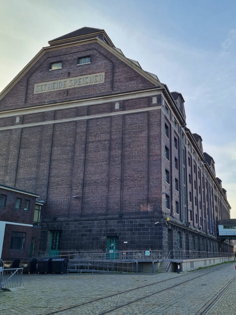Eine sehr große, alte Lagerhalle aus Backstein. An der rechten Seite viele Fenster. Die schmalere Gebäudeseite, auf die die Kamera gerichtet ist, hat nur oben zwei Fenster und darunter groß die Wörter "Getreide Speicher".