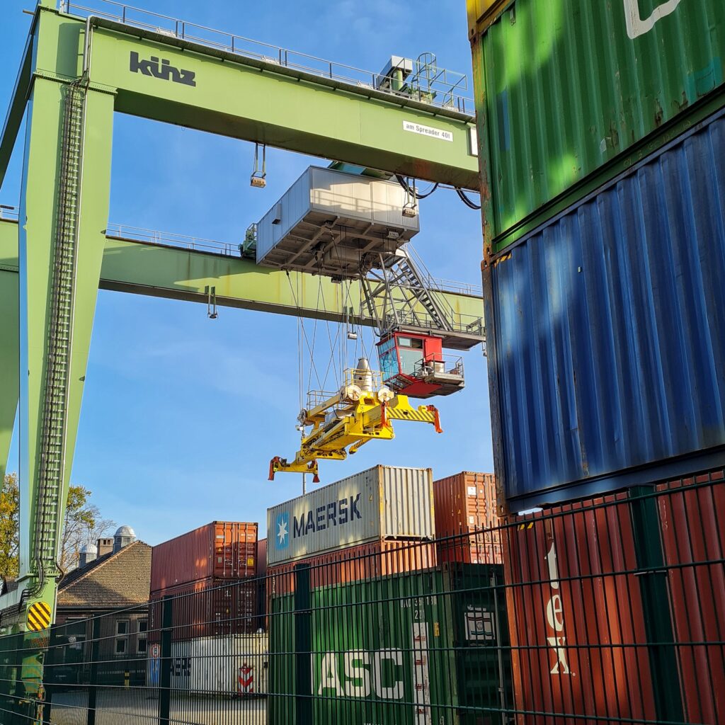 Stapel mit bunten Containern. An einer grünen Containerbrücke hängt ein gelber breiter Containerkran über einem grauen Container mit der Aufschrift "Maersk". Die rote verglaste Führerkabine der Containerbrücke hängt seitlich und etwas höher als der gelbe Kran.