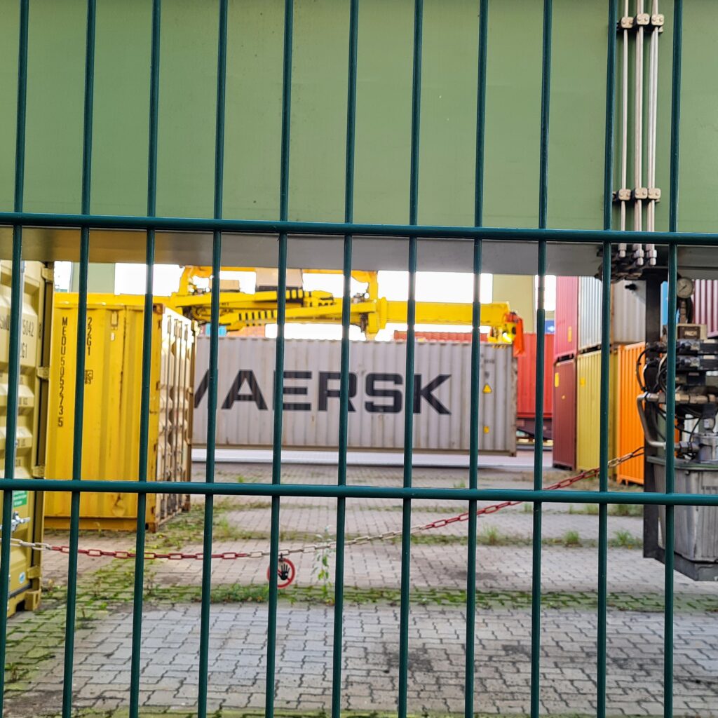 Blick durch einen Gitterzaum. In der Mitte zwischen bunten Containern wird durch einen gelben breiten Kran ein grauer Container mit der Aufschrift "MAERSK" auf den Boden gestellt.