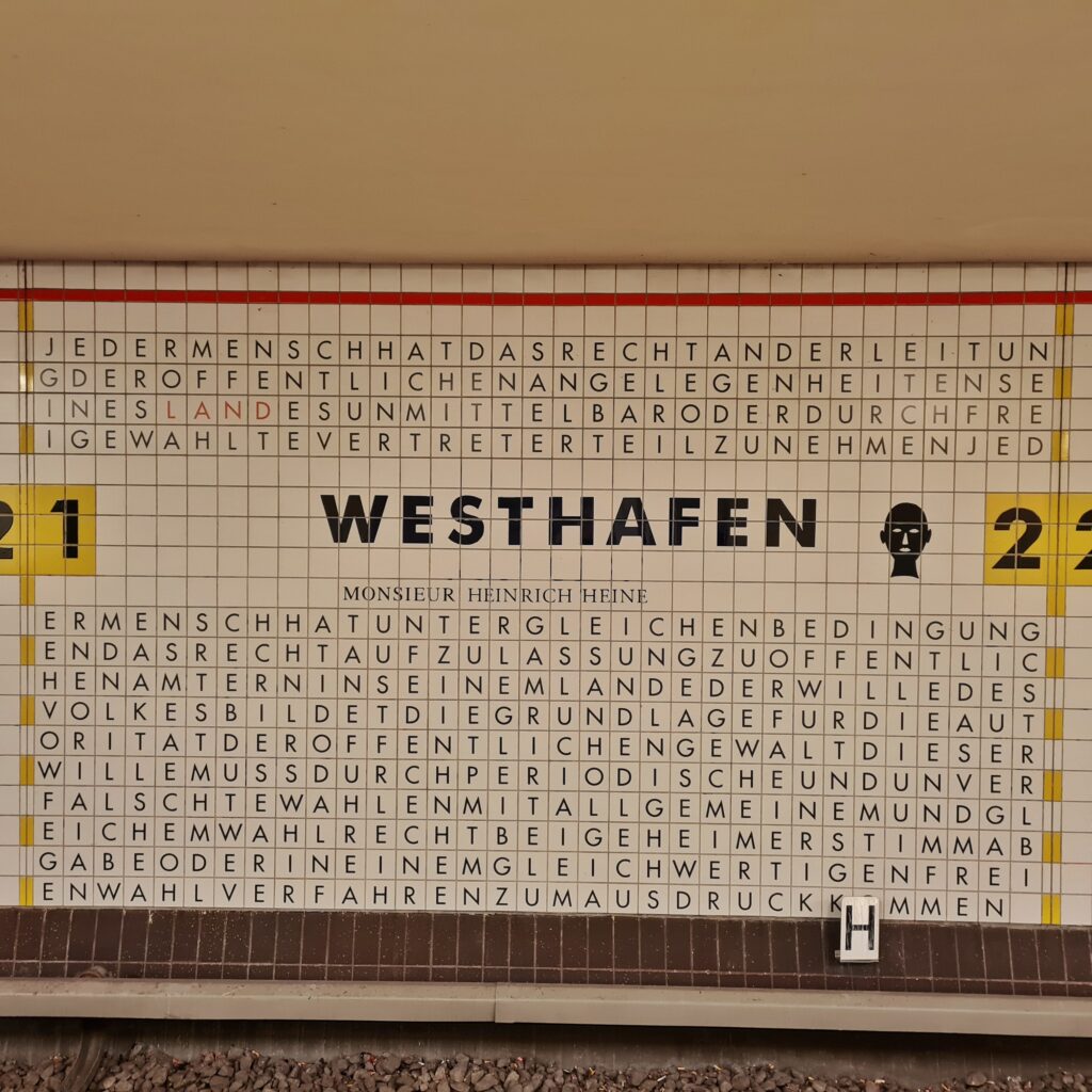 Weiße Fliesen an einer Wand. Auf jeder ein schwarzer Großbuchstabe. Da keine Lücken zwischen den Buchstaben sind, sieht es auf den ersten Blick aus, wie die Rätsel, bei denen man in Buchstabenwirrwarr Wörter finden muss. Wenn mann die Buchstaben aber nach und nach liest und gedanklich die Lücken einfügt, erkennt man, dass es sich um sinnvollen Text handelt. In der oberen Hälfte steht in großer Schrift Westhafen. Darunter: Monsieur Heinrich Heine. Rechts daneben due schwarze Zeichnung eines Kopfes. Links gelbe Fliesen mit der großen Zahl 21, rechts 22.