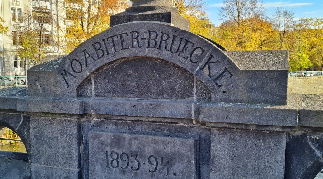 Ein Steinelement auf einer Brücke. Oben ein kronenförmiges Objekt. Darunter ein Bogen mit der Aufschrift: Moabiter-Brücke. Unten die Aufschrift: 1893 - 94. Im Hintergrund mehrstöckige Häuser und bunt gefärbte Bäume.