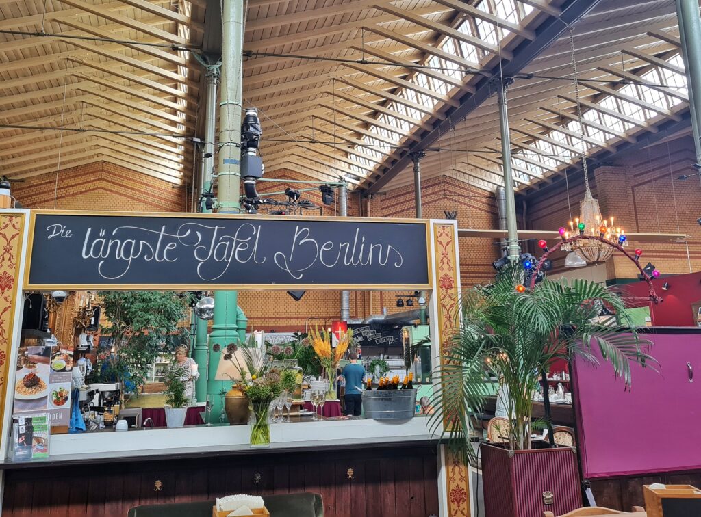 Im Vordergrund ein Tresen mit einer weißen Platte. Darüber eine Tafel, auf der mit Kreide geschrieben steht: Die längste Tafel Berlins. Im Hintergrund Tische und Stühle, Geschirr, Blumenschmuck, einige Menschen, Stände mit Essen, rechts oben ein Kronleuchter. Oben ist gut die Dachkonstruktion der Halle mit seitlichen Fensterreihen zu sehen.