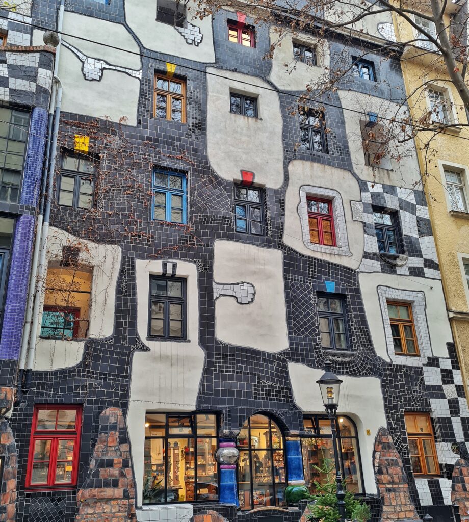 Eine mehrstöckige Hausfassade. Teilweise ist sie mit schwarzen Fliesen in unregelmäßigen Formen verkleidet. Um die Fenster herum sind weißliche Flächen mit unregelmäßigen Kanten. Die Fenster sind Sprossenfenster in unterschiedlichen Größen. Ihre Rahmen sind in bunten Farben bemalt. Im Erdgeschoss ist ein Laden.