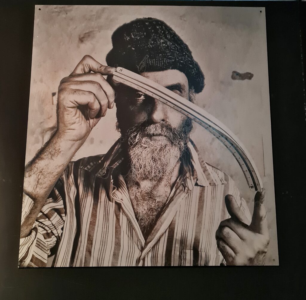 Ein Foto von Hundertwasser, der zwischen beiden Händen ein gebogenes Teil hält. Er hat ein gestreiftes Hemd an und auf dem Kopf eine Wollmütze.
