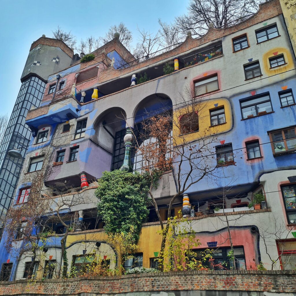 Ein mehrstöckiges Haus, das in unregelmäßgen Flächen bunt bemalt und teilweise begrünt ist, vor allem im die Fenster herum. Es gibt mehrere von außen nicht einsehbare Balkone. In der Mitte des Gebäudes sind zwei hohe Rundbögen mit einem Pfeiler dazwischen. Dahinter eine große Fensterfläche. 