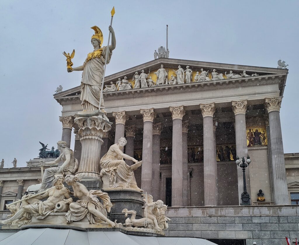 Das tempelartige Gebäude mit den Säule von vorne. Davor ein Denkmal: auf einer weißen Steinsäule steht eine Frauenfigur mit einem Speer in der einen Hand und einer Engelsfigur auf der ausgestreckten anderen Hand. Die mützenartige Kopfbedeckung, der Brustpanzer, die Speerspitze und die Engelsfigur sind goldfarben. Am Fuß der Säule vier weitere sitzende Figuren: eine Frau mit einer Steintafel in der Hand, eine mit einem Schwert, eine halbnackte Frau, die sich einem halbnackten Mann mit gelockten Haaren und Bart zuwendet.