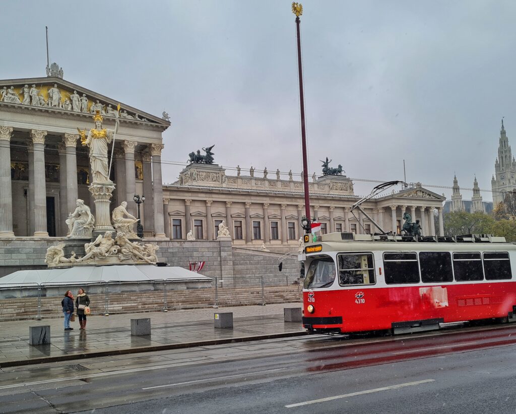 Hinter einer Straße, auf der eine rote Straßenbahn fährt, ein länglicher Gebäudekomplex. Links ein tempelartiges Gebäude mit hohen Säulen. Daneben ein längliches Gebäude mit nebeneinander liegenden Fenstern. Auf dem Dach mehrere Steinstatuen und an den beiden Seiten auf Podesten jeweils eine Quadriga aus Metall.