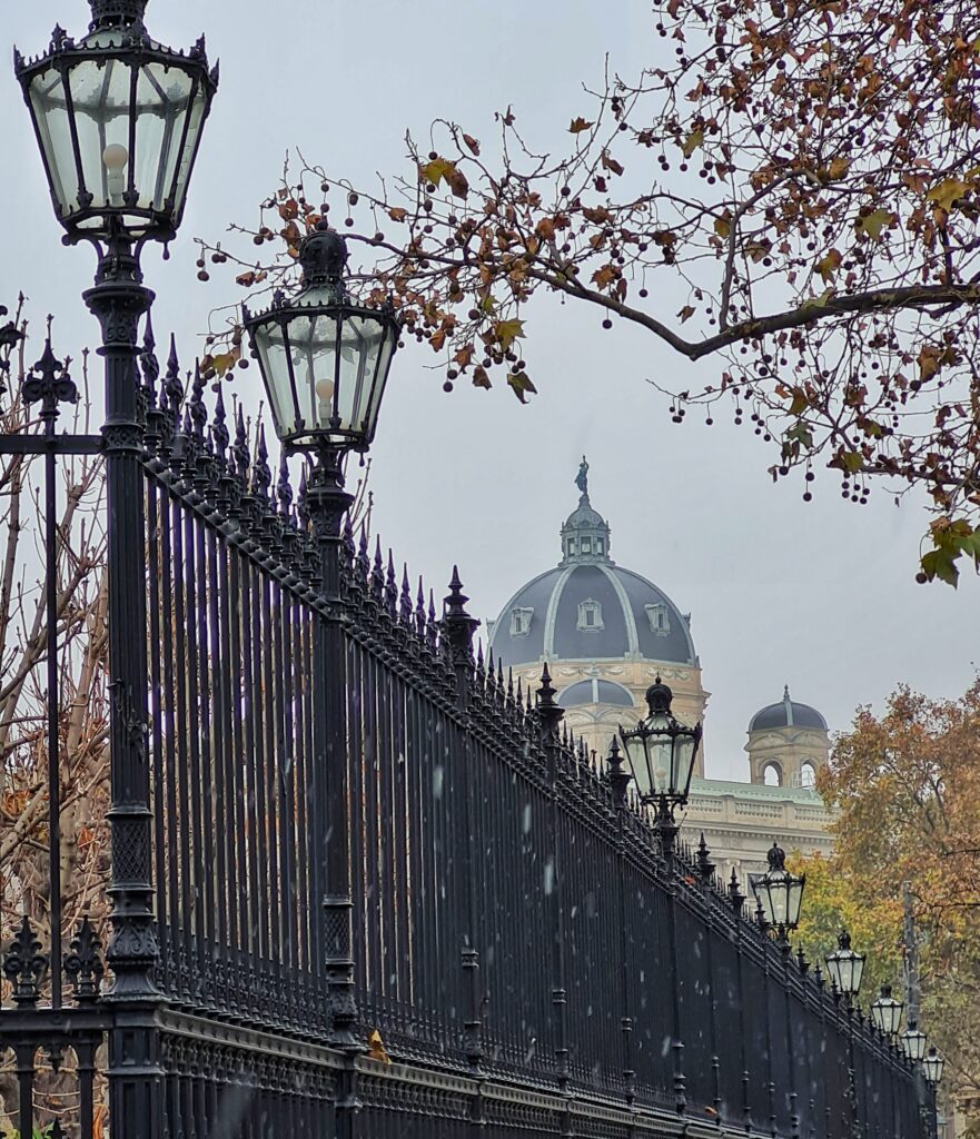 Blick über einen Metallzaun. Die Metallstäbe stehen eng nebeneinander. Oben läuft der Zaun in Spitzen aus. Am Zaun zwei altmodische Laternen. Im Hintergrund eine große Gebäudekuppel.