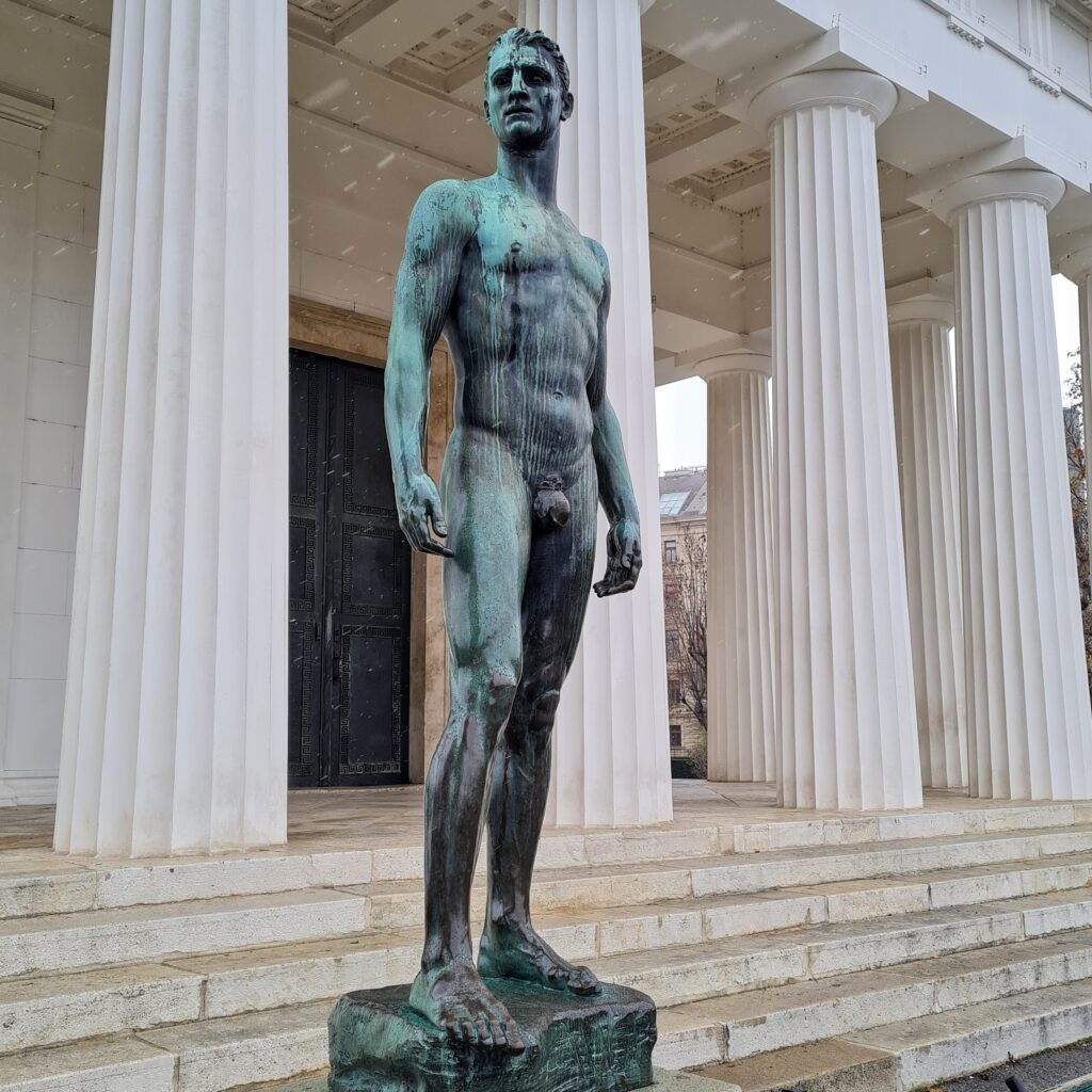 Teil eines Gebäudes mit hellen geriffelten Säulen und einigen Treppenstufen. Davor die Metallstatue eines nackten jungen Mannes mit sportlichem Körperbau.