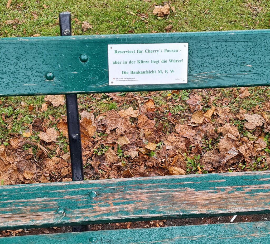Ein Teil einer grünen, teilweise abgeblätterten Bank. An der Lehne ist ein weißes Schild angebracht, auf dem steht: "Reserviert für Cherry's Pausen - aber in der Kürze liegt die Würze! Die Bankaufsicht M, P, W".