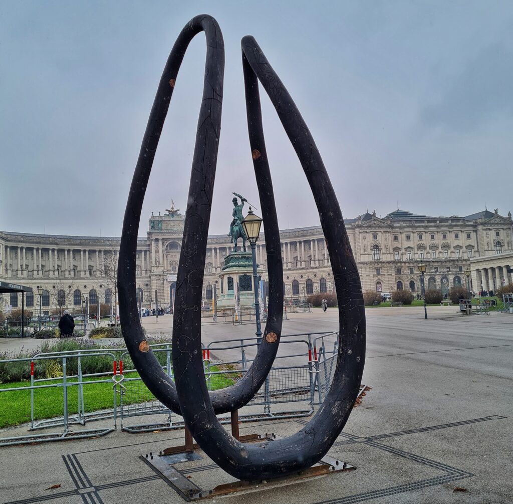 Blick auf die Hofburg und den Heldenplatz mit einem großen Reiterstandbild auf einem rechteckigen Podest. Im Vordergrund ein Denkmal in Form einer mehrfach gebogenen Holzschleife.